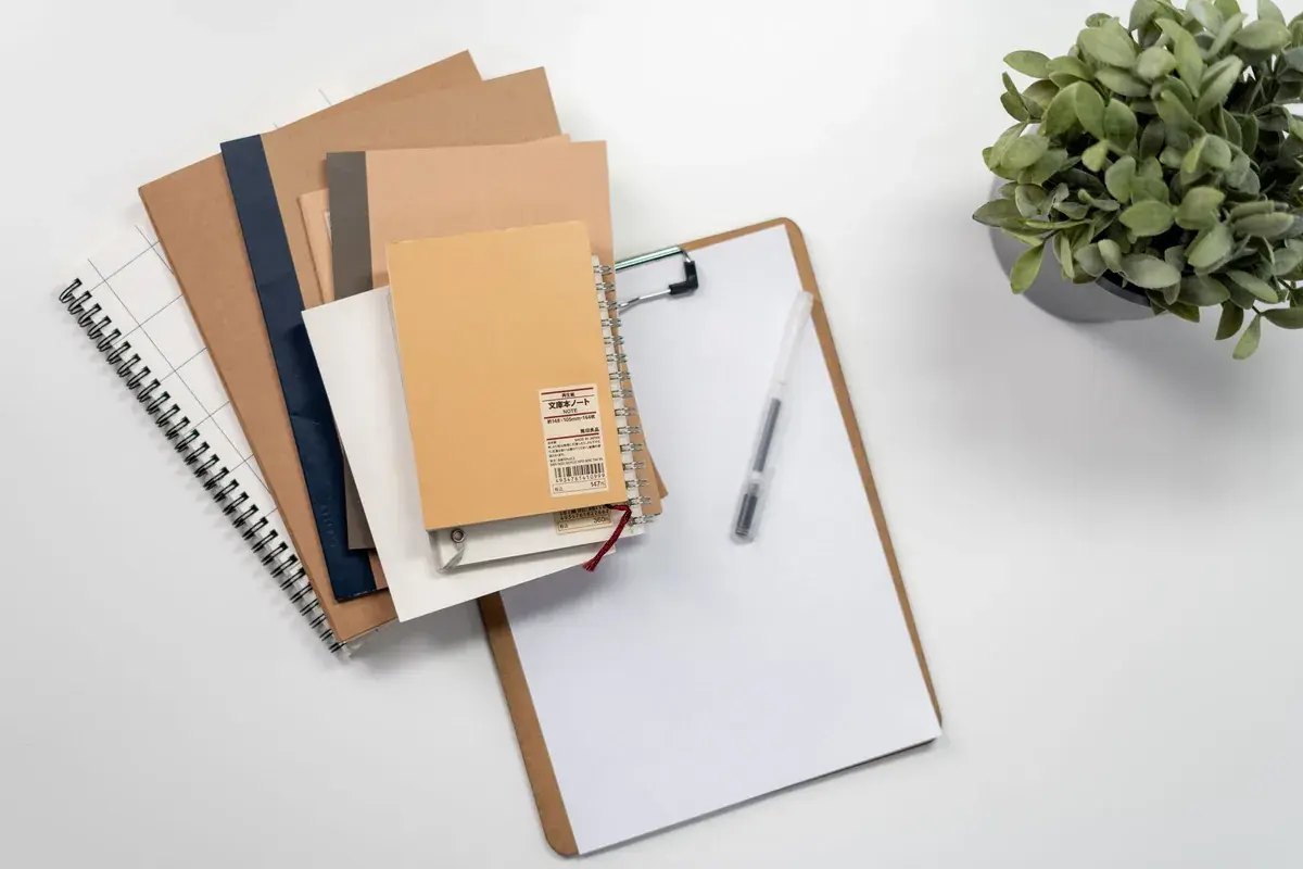 A tidy desk seen from above: notebooks, a clipboard, a pen and a small plant.