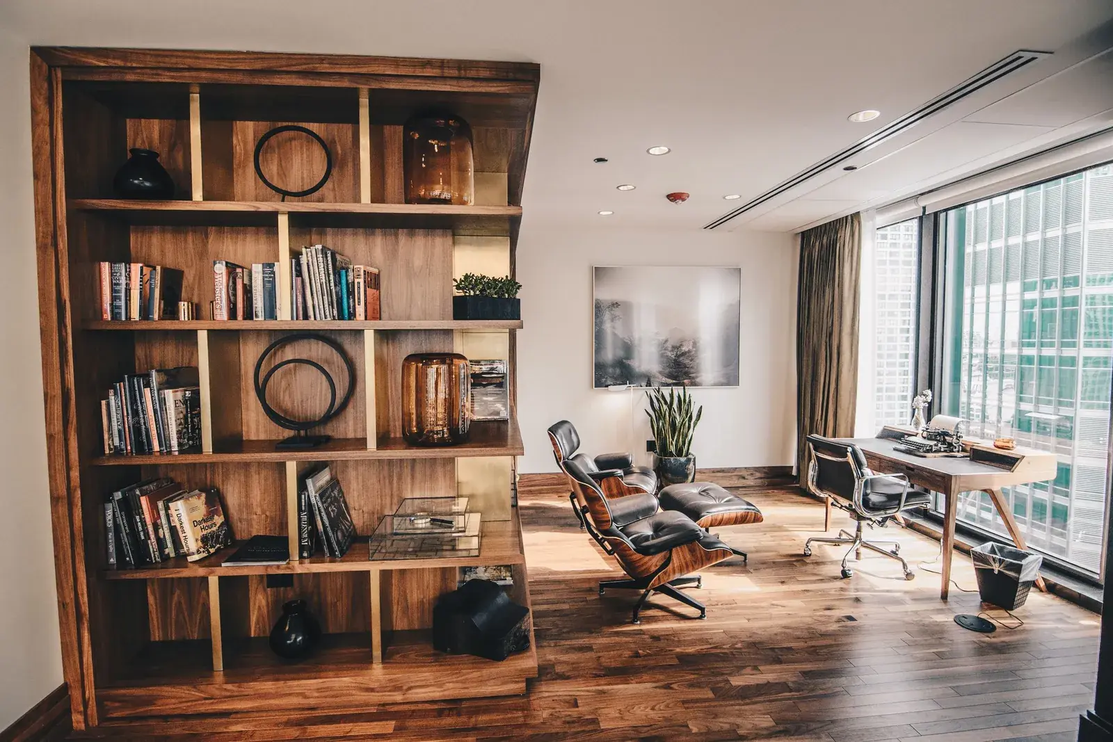 A quiet office with a wooden bookcase, an armchair and a long desk by the window.