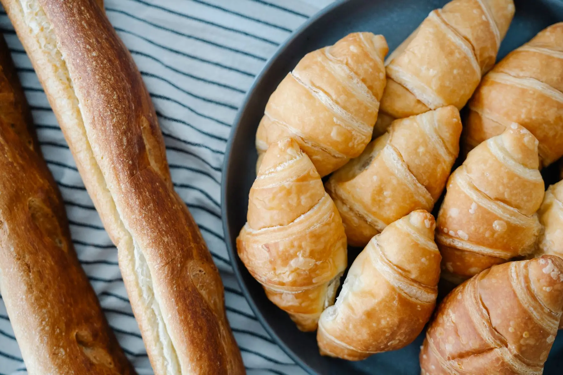 Golden croissants on a dark plate beside a baguette, on a striped cloth.