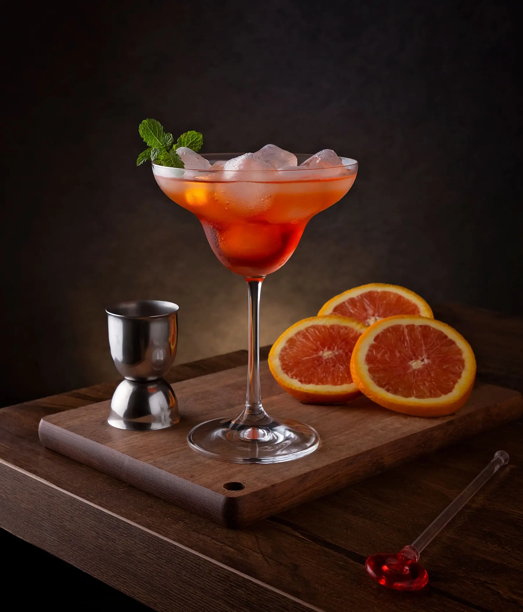 A coupe of orange cocktail beside a jigger and sliced oranges on a wooden board.