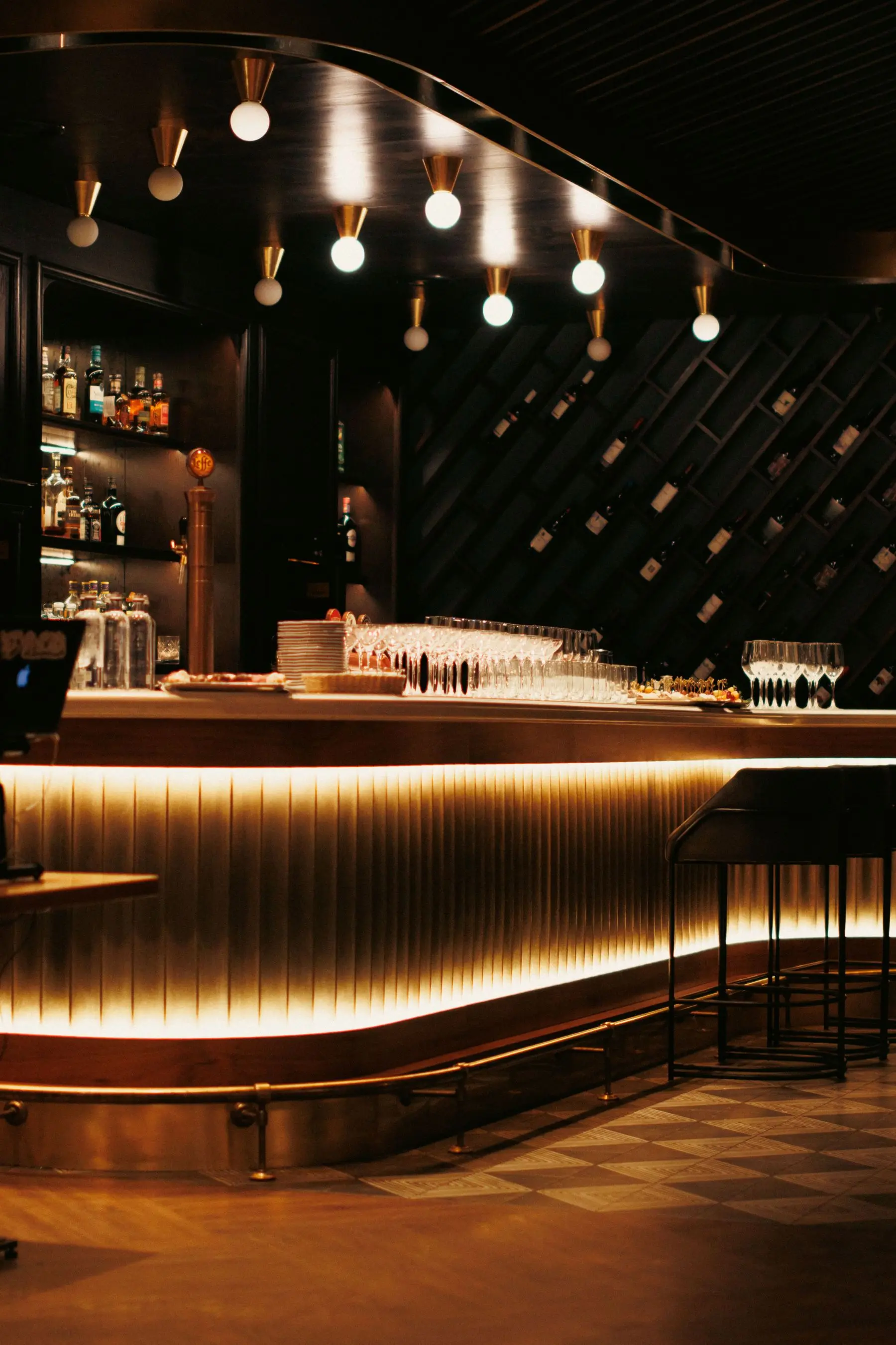 A sleek bar counter lit from below, glasses lined up and bottles on dark shelves behind.