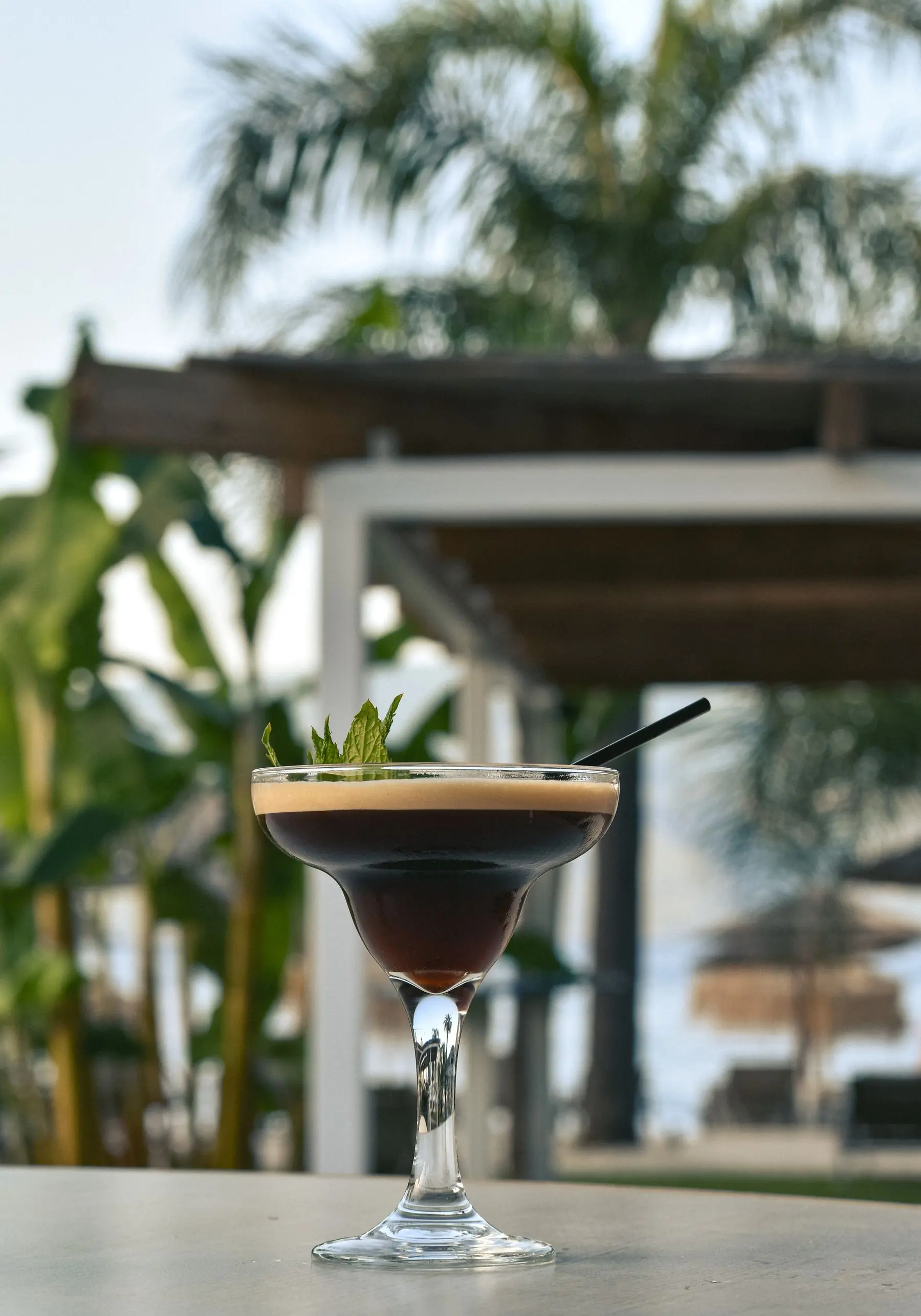 An espresso martini in a coupe, a mint leaf on its foam, a terrace blurred behind.