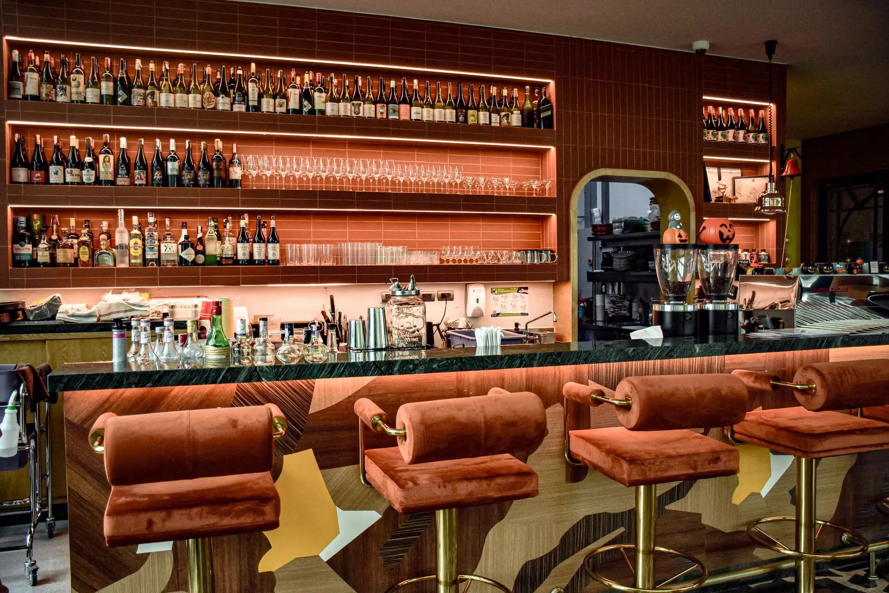 Rust-coloured velvet bar stools at a counter under a wall of backlit bottle shelves.