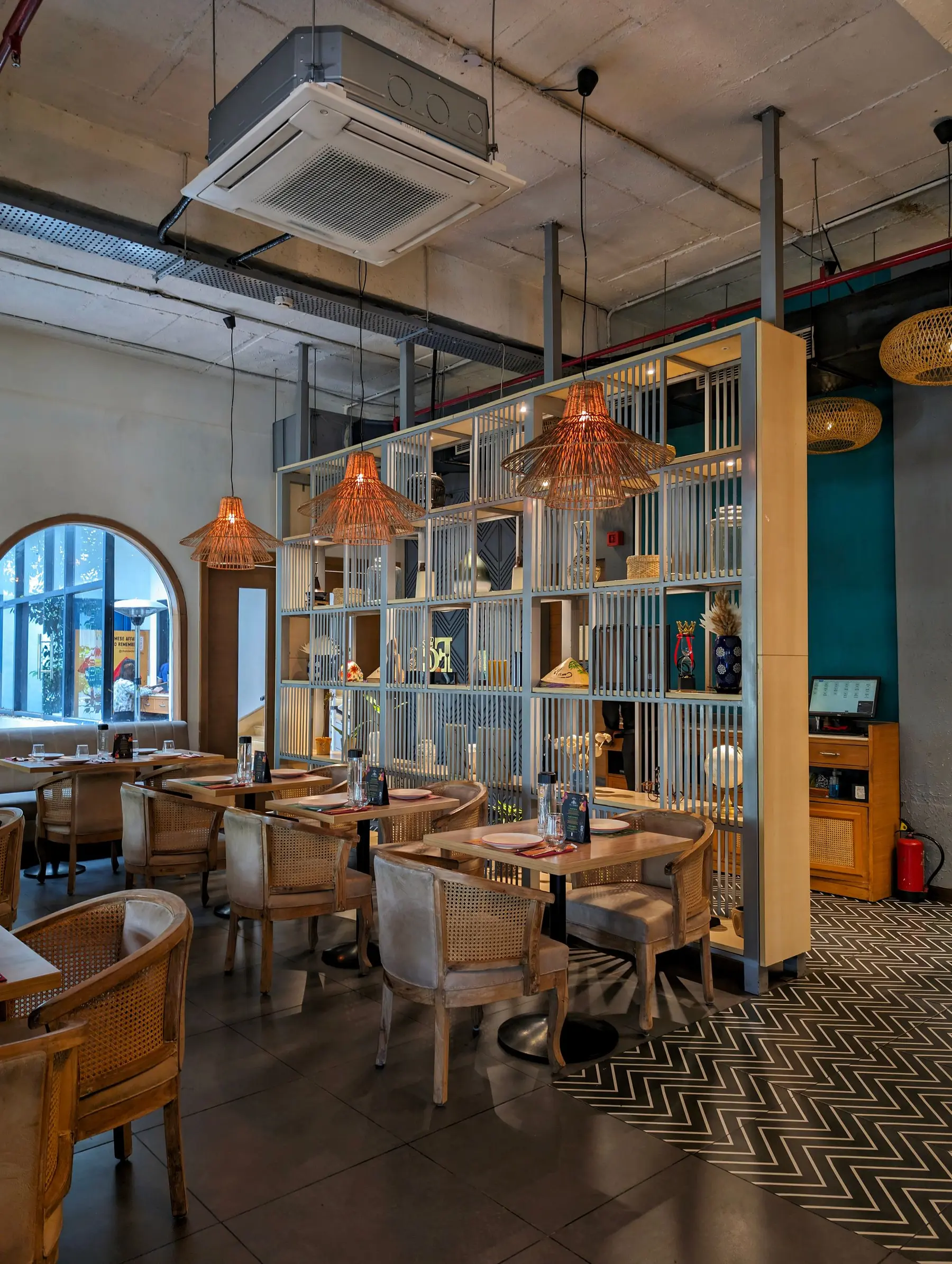 A café with cane chairs, woven pendant lamps and a tall shelf of plants and books.