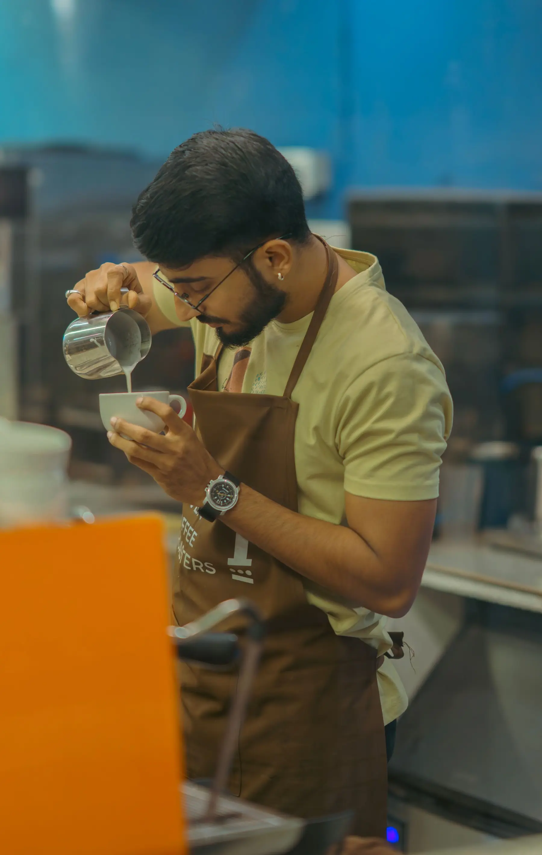 A barista in an apron pours milk into a cup, steam rising.