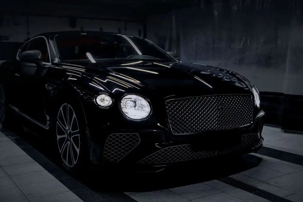 A black grand tourer, three-quarter front, its headlamps lit in a dark garage.
