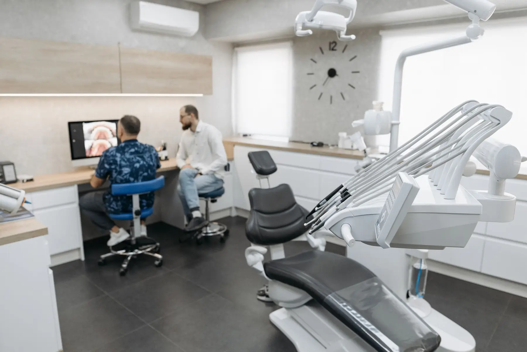 A bright, modern dental clinic with a chair, wall clock and two people talking at the desk.