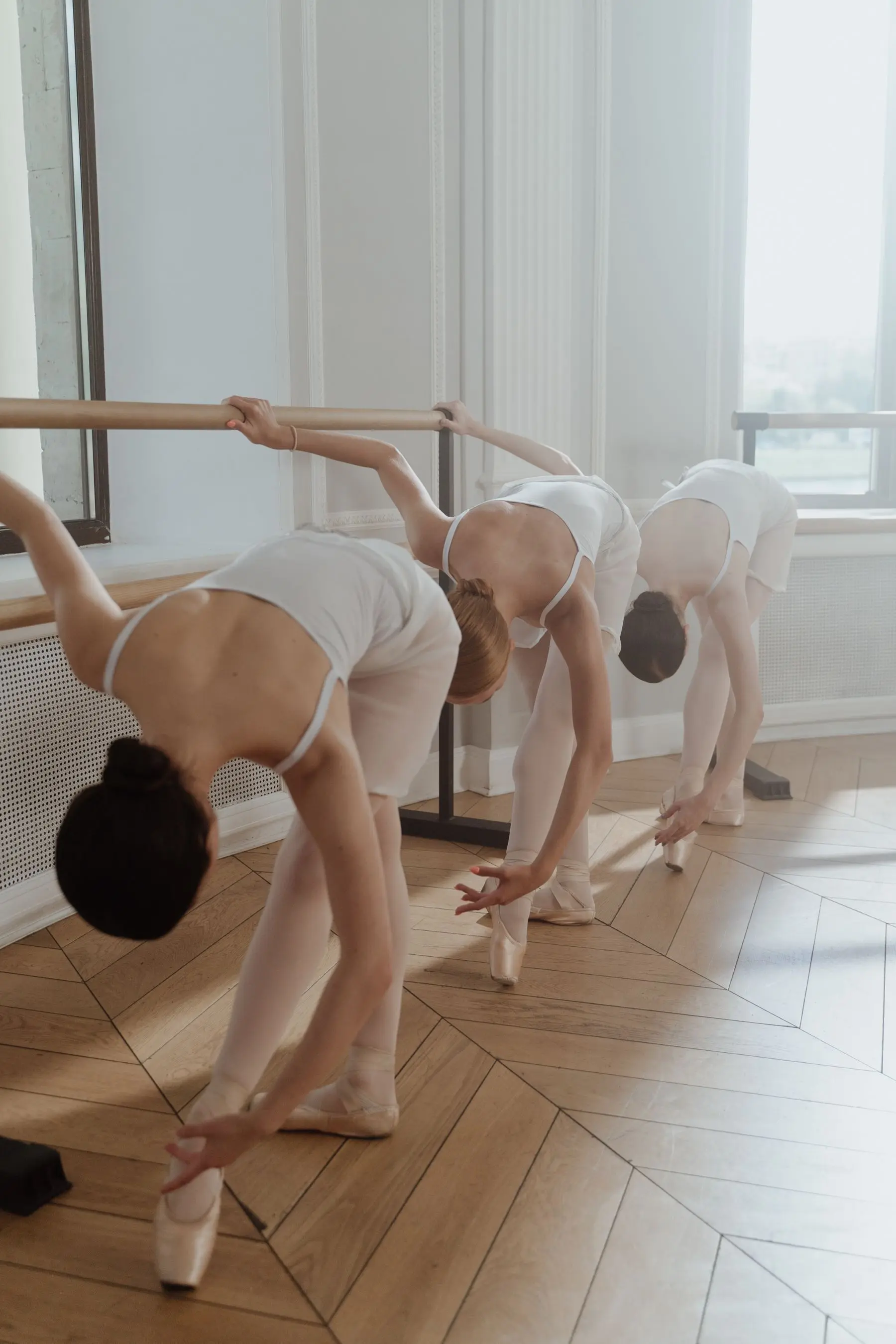 Dancers in white bend deeply backwards at a ballet barre in a bright studio.