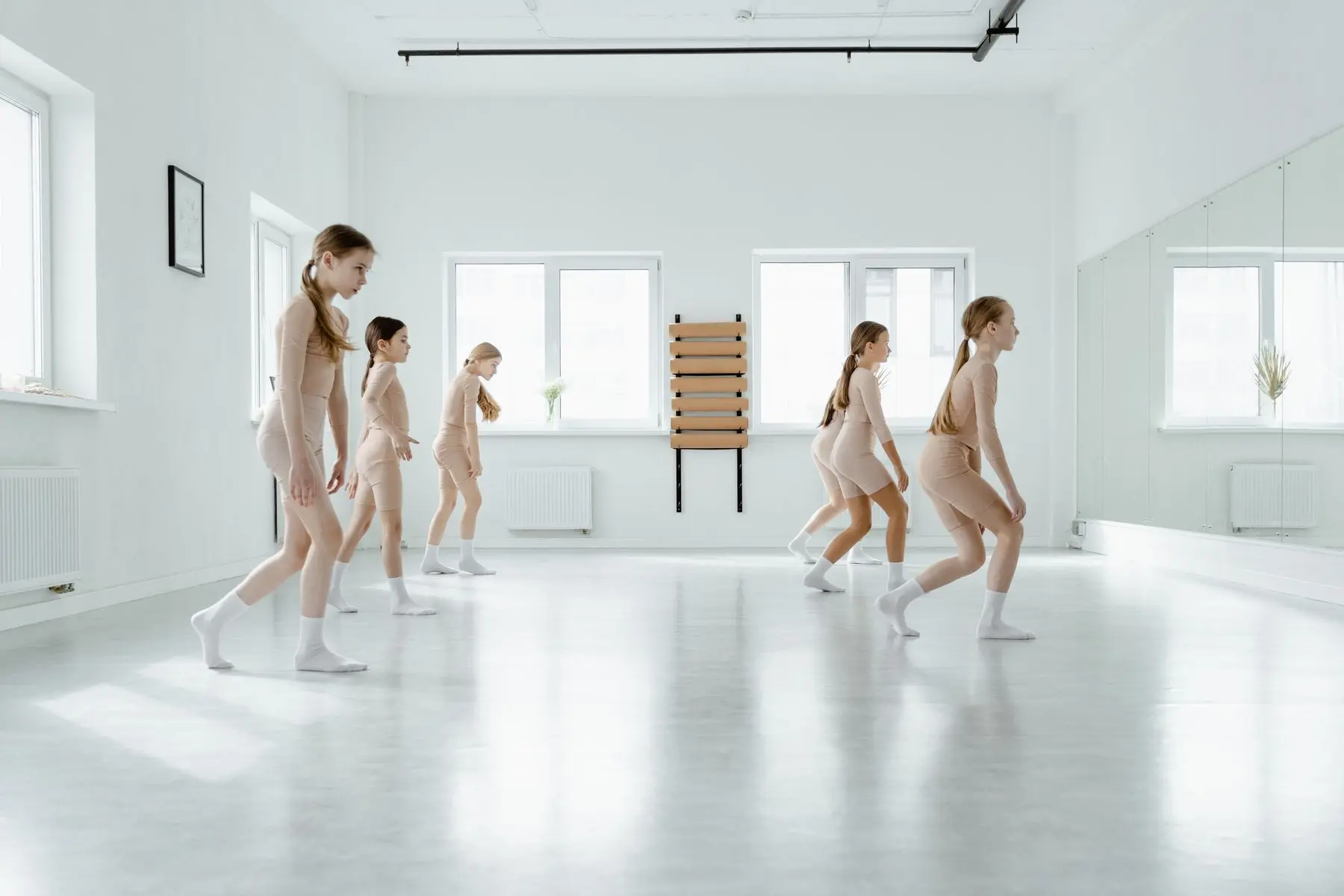 Young dancers in a row rehearse a crouching step across a bright, white studio.
