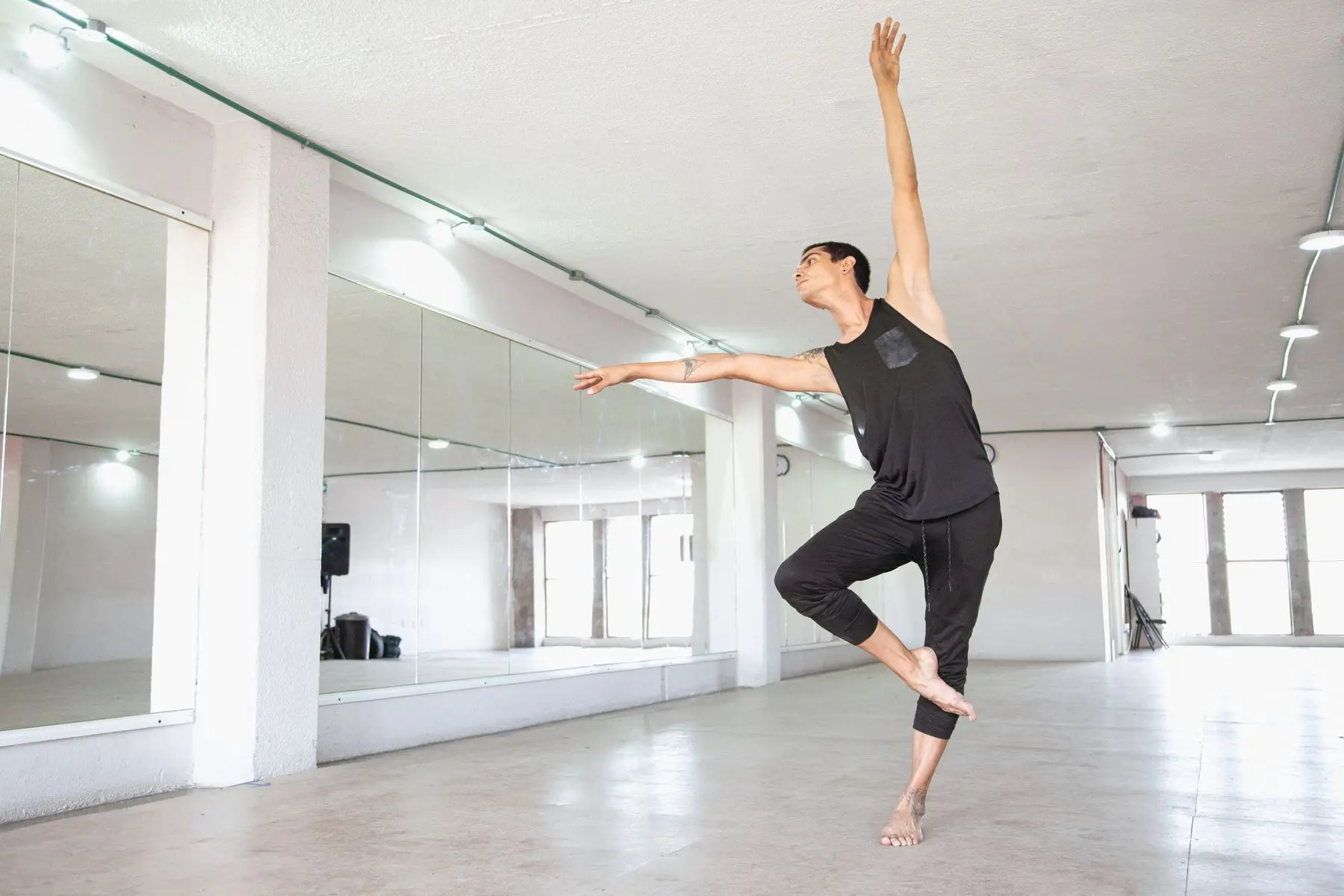 A dancer in black balances on one leg with an arm raised in front of a mirrored wall.