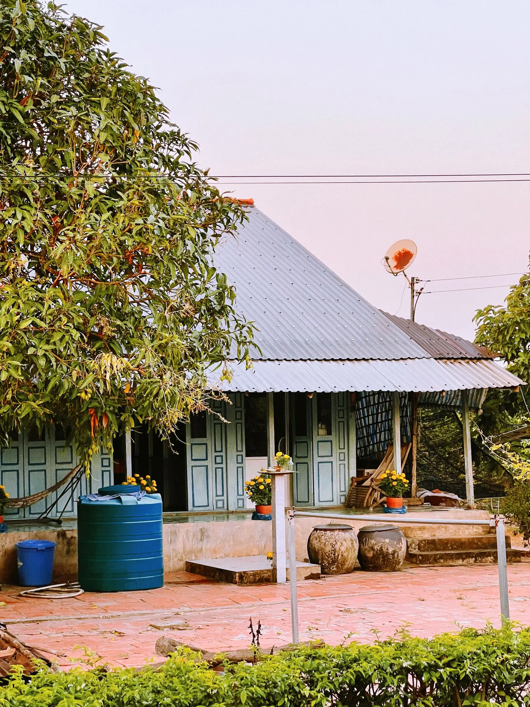 The house's veranda under a corrugated roof, with a hammock, pots and a water tank in the evening light.