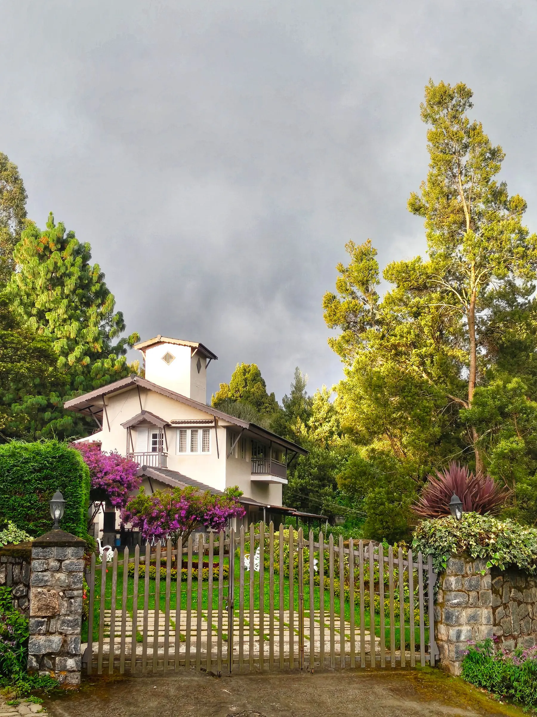 The two-storey house among tall trees and flowering shrubs, seen from the path.