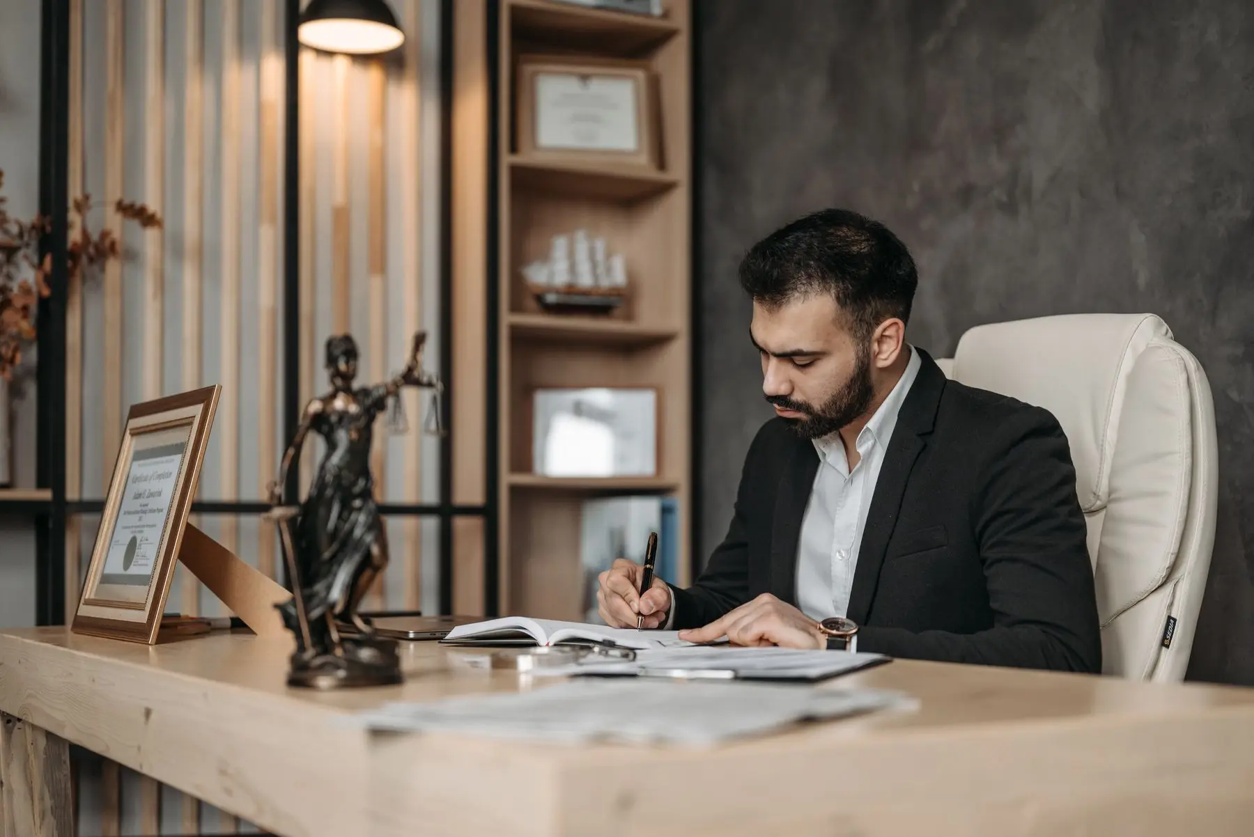 A bearded lawyer in a black suit writes notes at a desk, a Lady Justice statuette beside him.