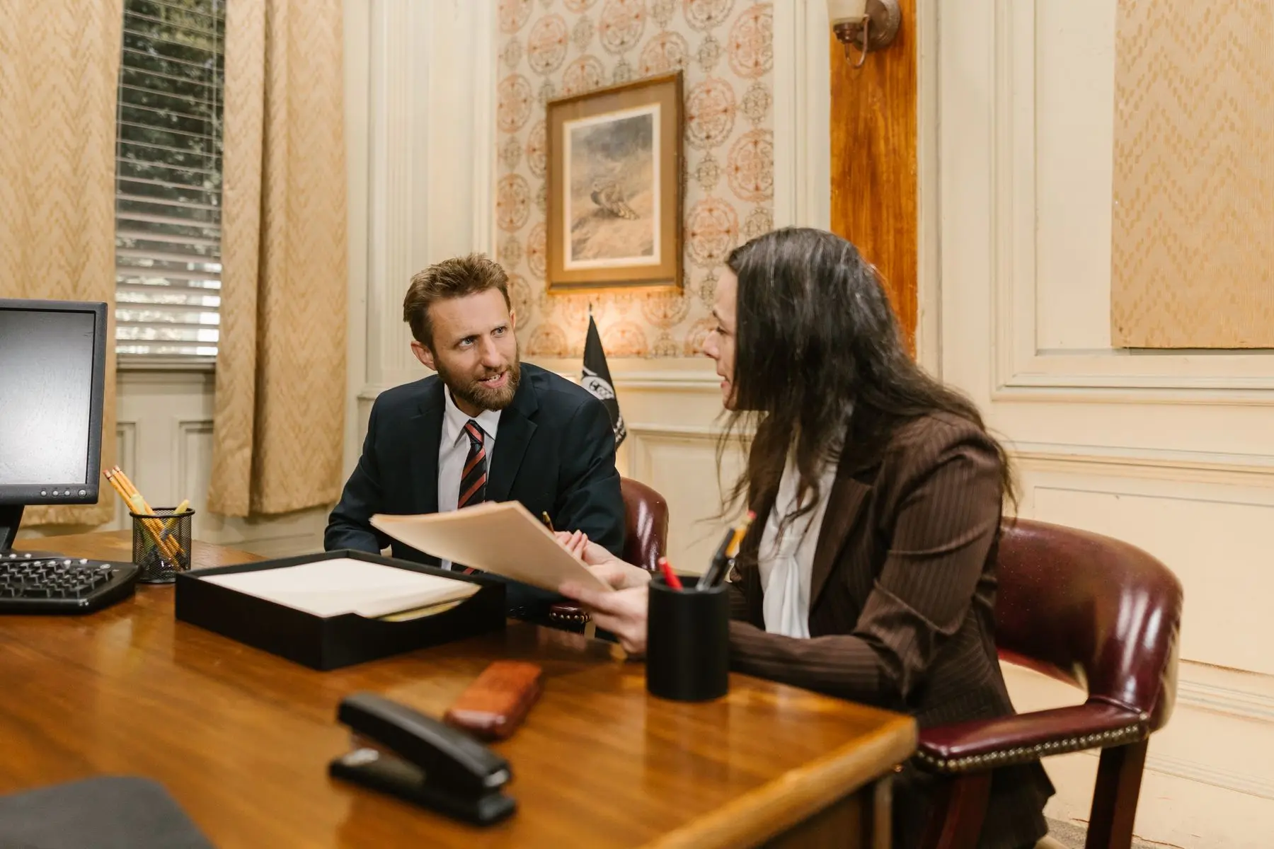A lawyer and a client talk across a desk in a wood-panelled office with framed pictures.