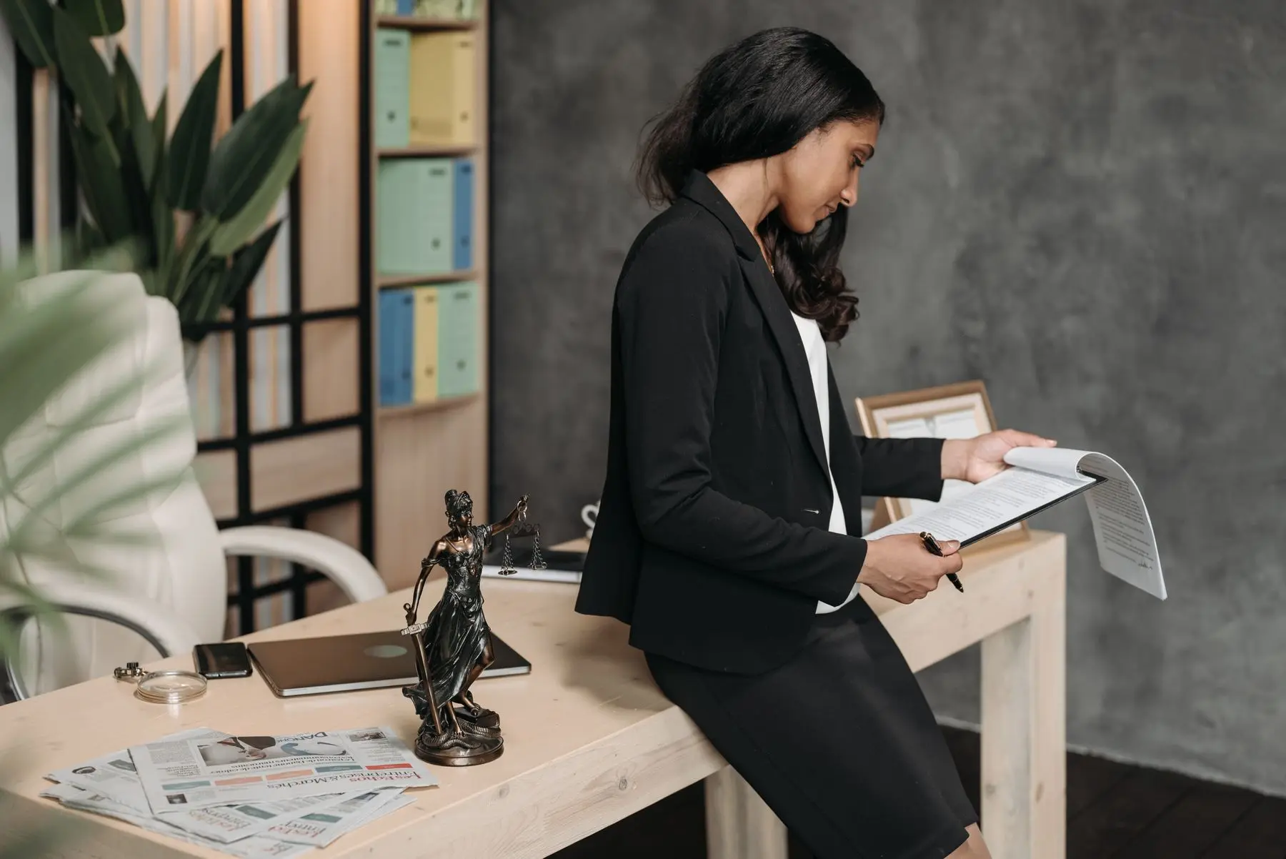 A woman lawyer in a black suit leans on a desk, reading a document, a statuette of Lady Justice beside her.