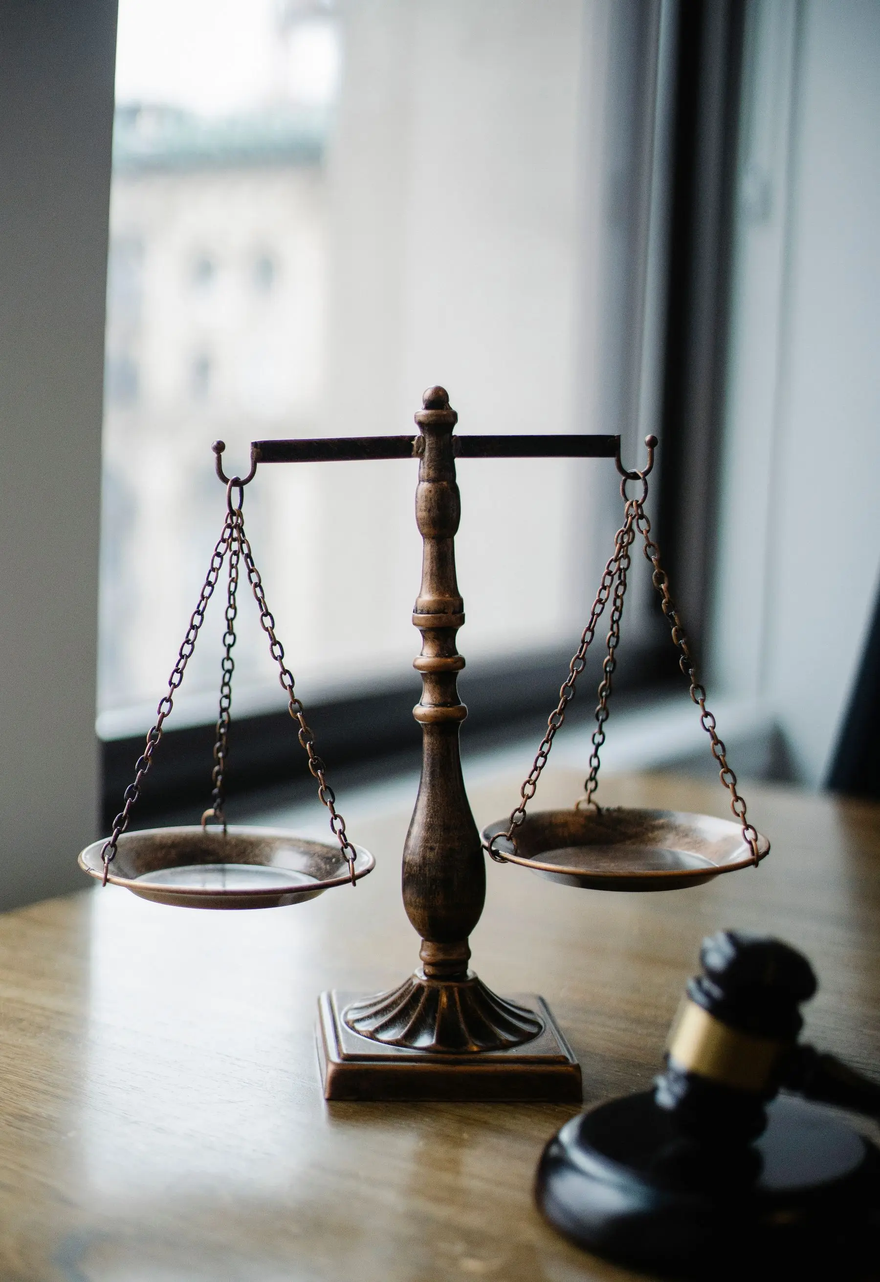 Antique brass scales of justice on a desk by a window, a gavel beside them.