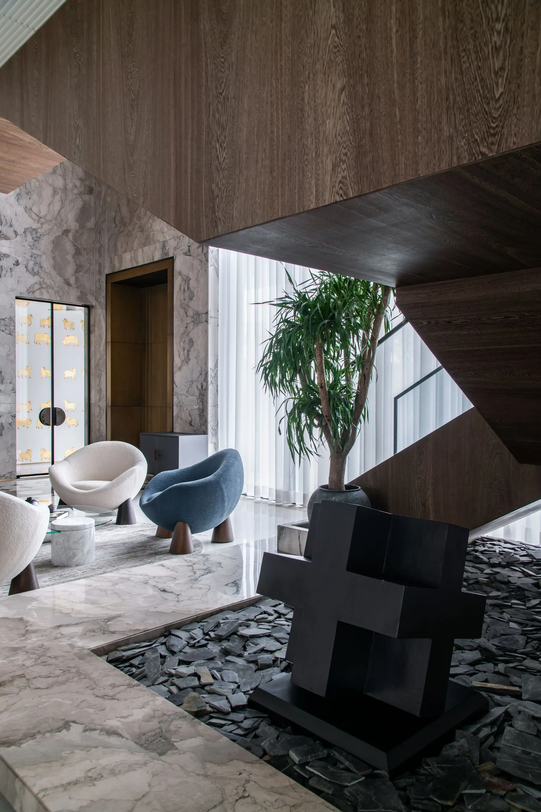 A double-height living space with sculptural white and blue chairs, a tall plant and marble walls.