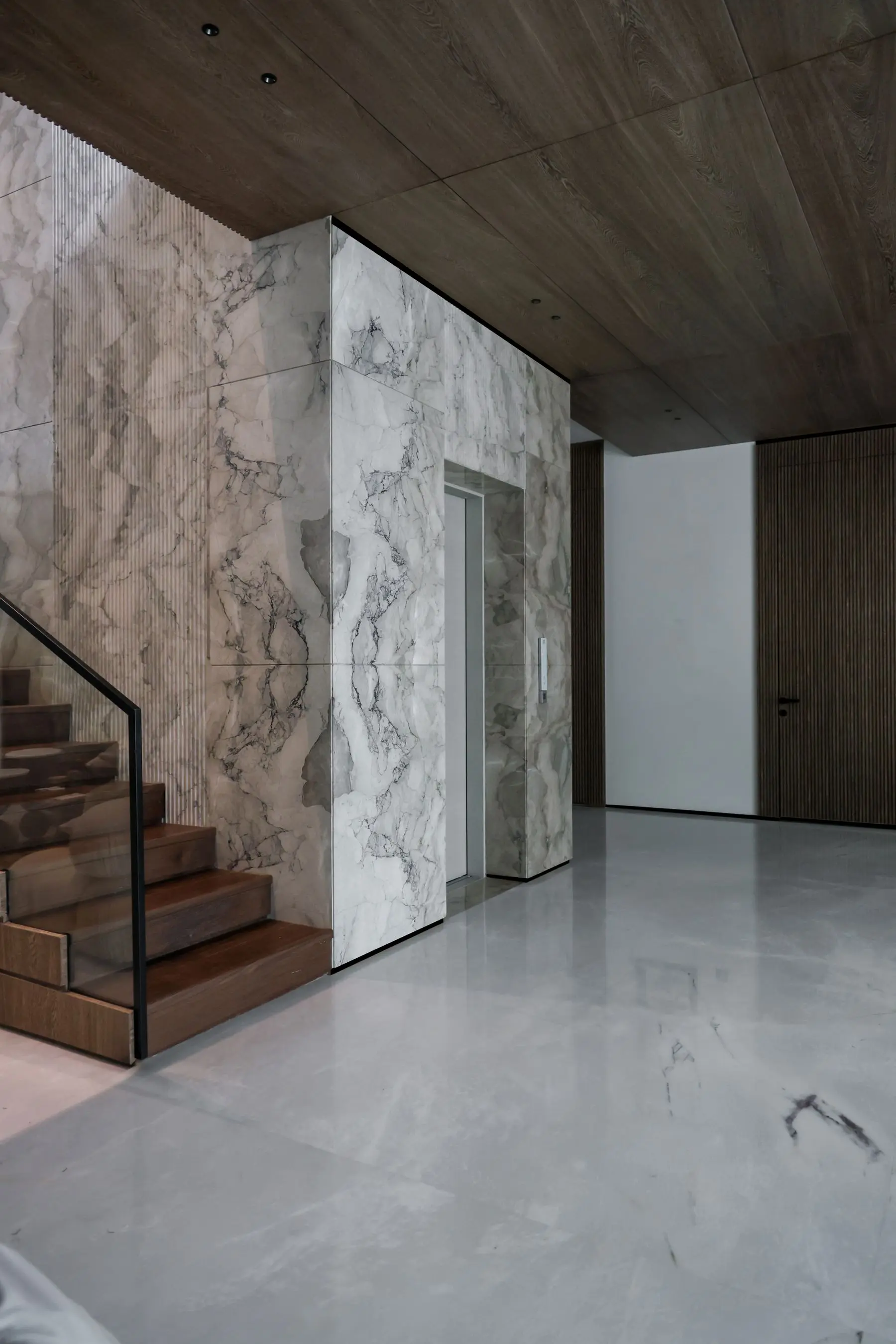 A stairwell clad in grey marble with a timber staircase and a glass balustrade.