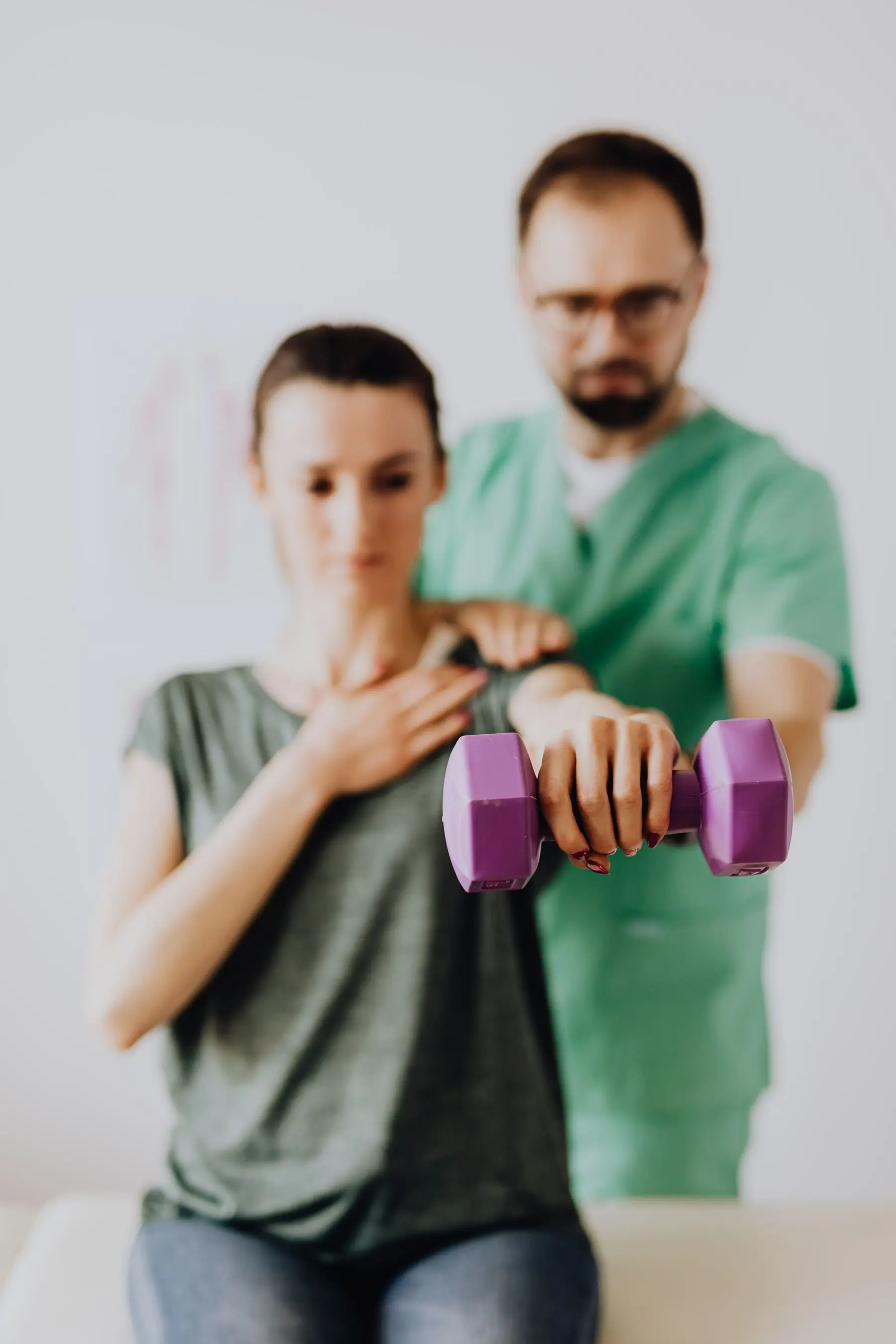 A therapist in green scrubs steadies a woman's shoulder as she lifts a small purple dumbbell.
