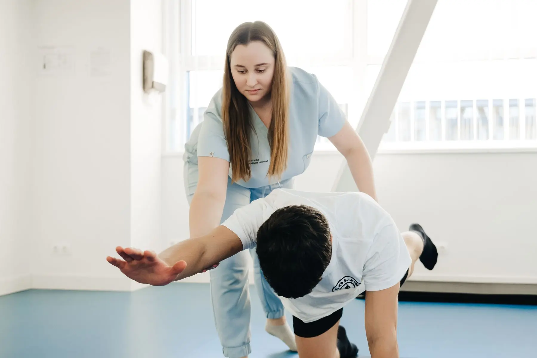 A physiotherapist in pale blue scrubs guides a man through a floor exercise on a mat in a bright clinic.