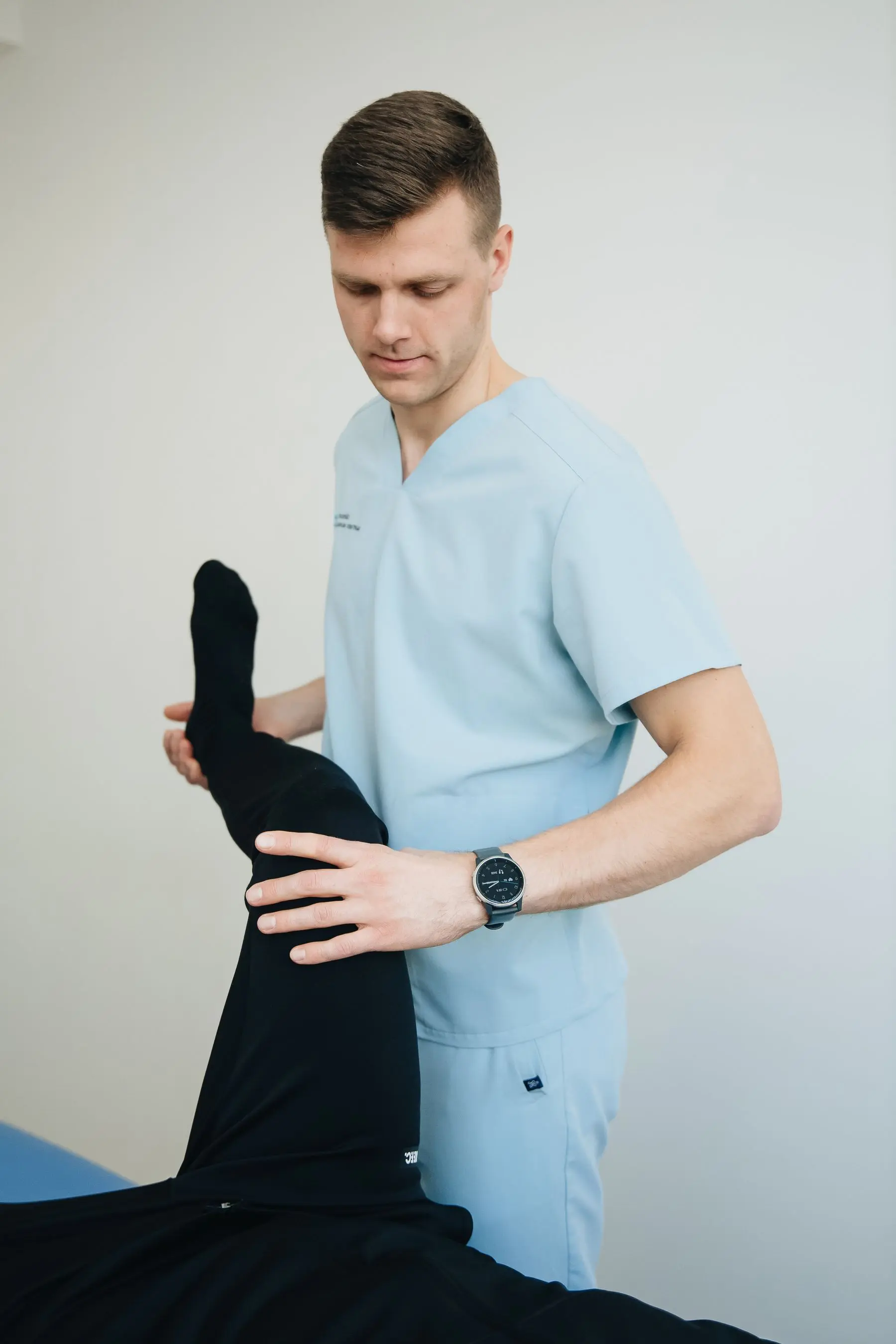 A physiotherapist in pale blue scrubs lifts a patient's straightened leg for a hamstring stretch.