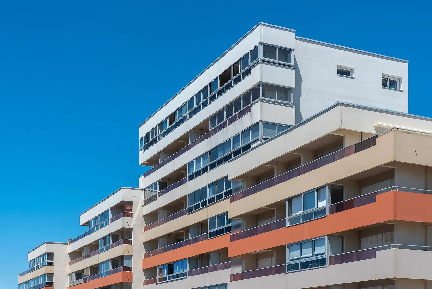 A low-angle view of a modern apartment block with long balconies and orange panels against a blue sky.