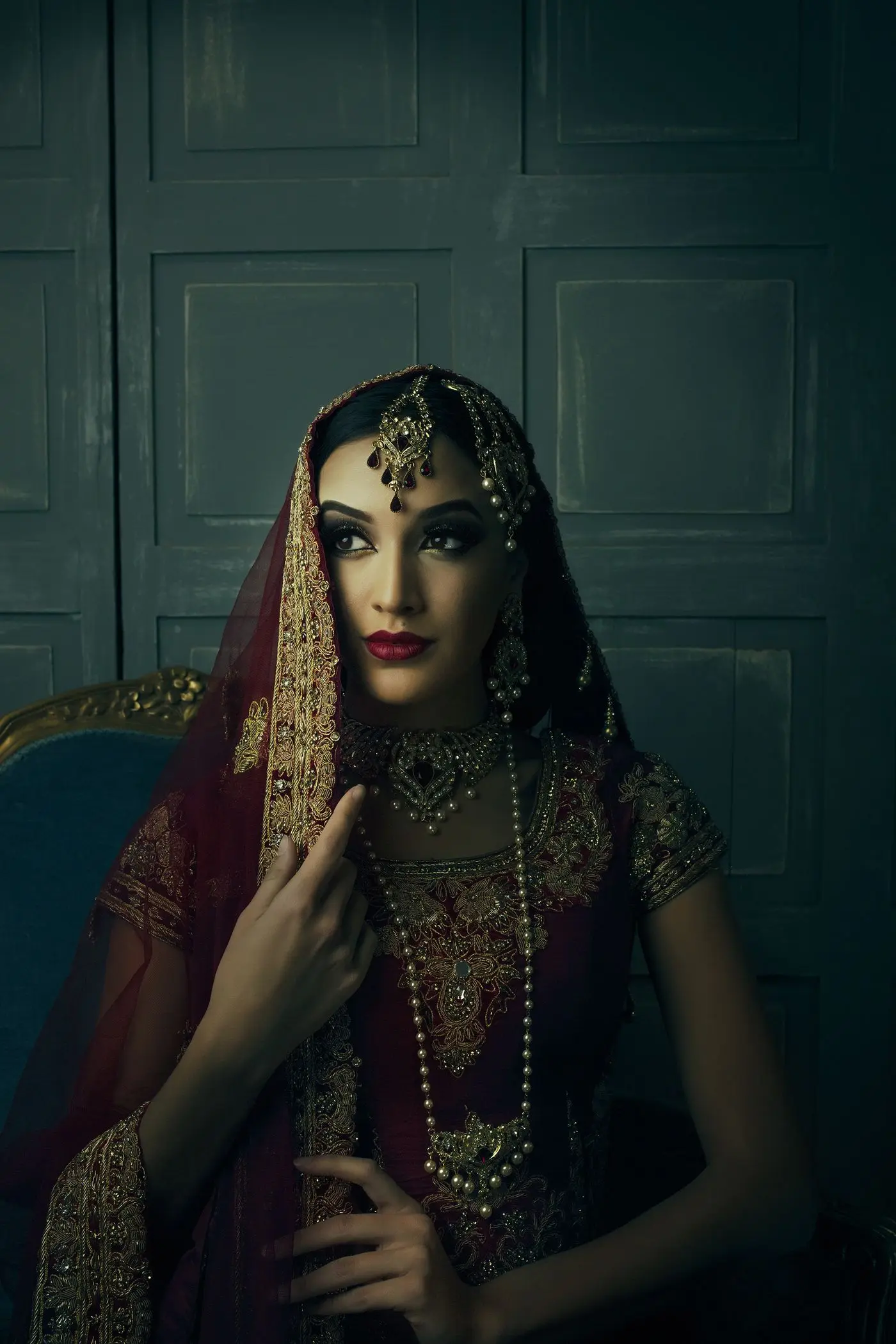 A woman in a red veil and gold jewellery, lit moodily against a dark teal wall.