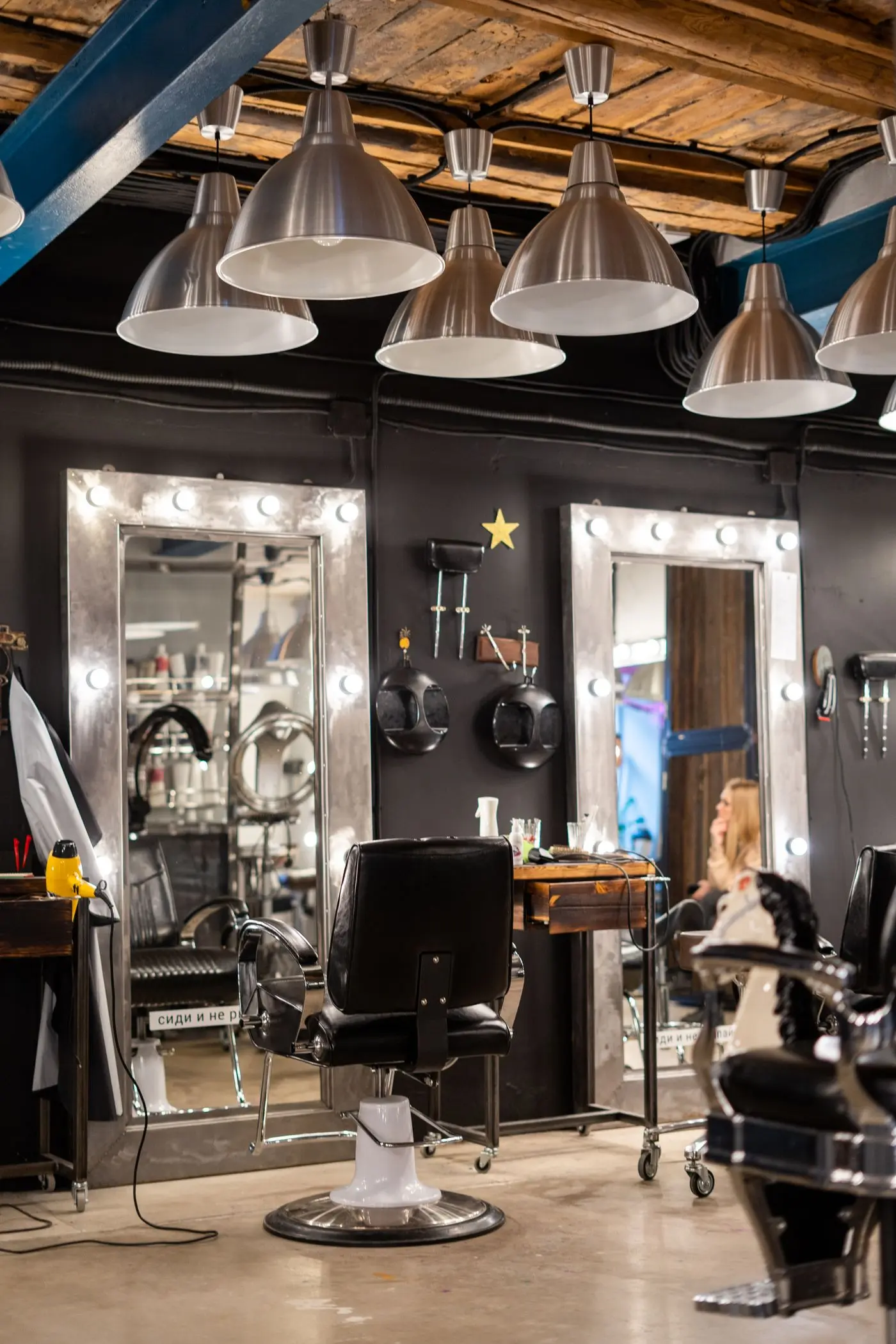 A dark salon with two mirrors framed by round bulbs and black styling chairs.