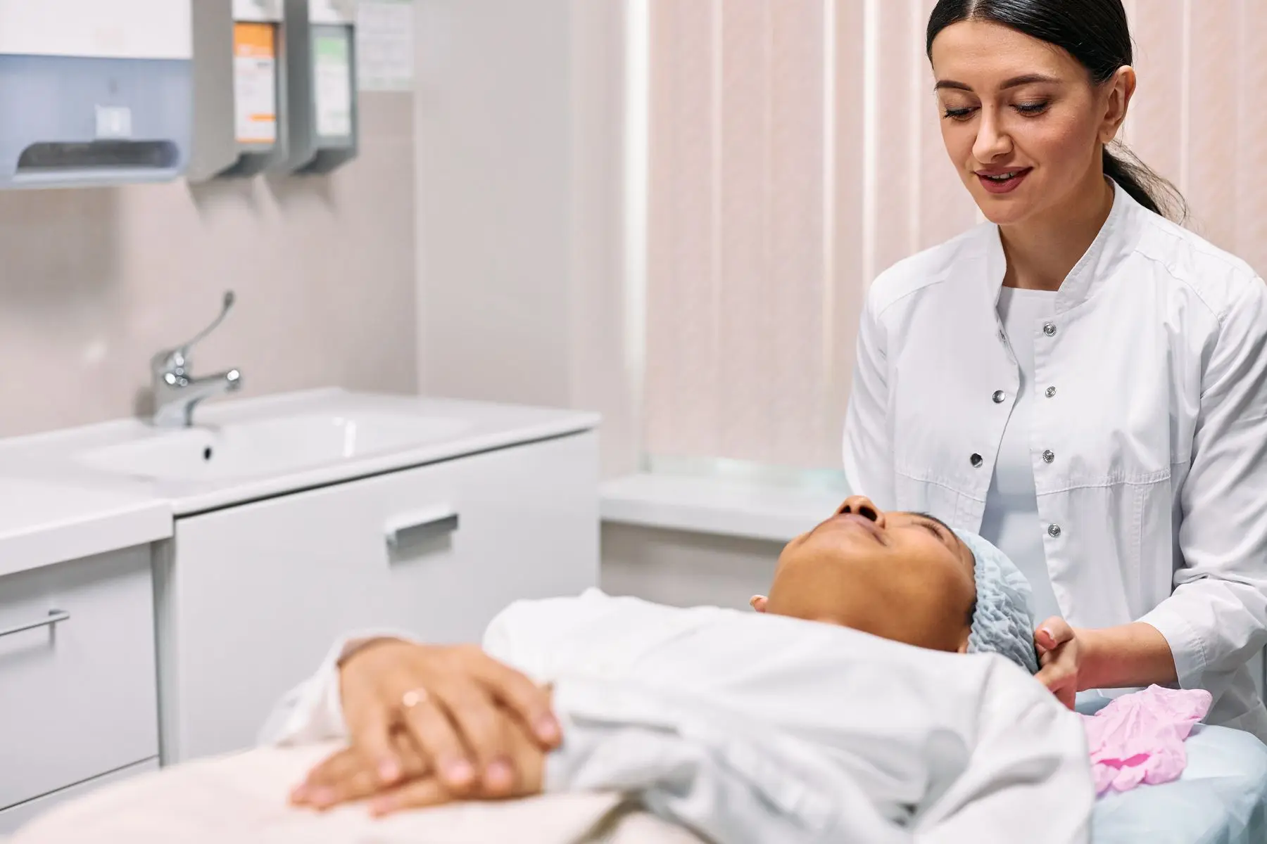 A beautician in white smiles while a client rests on a treatment bed.