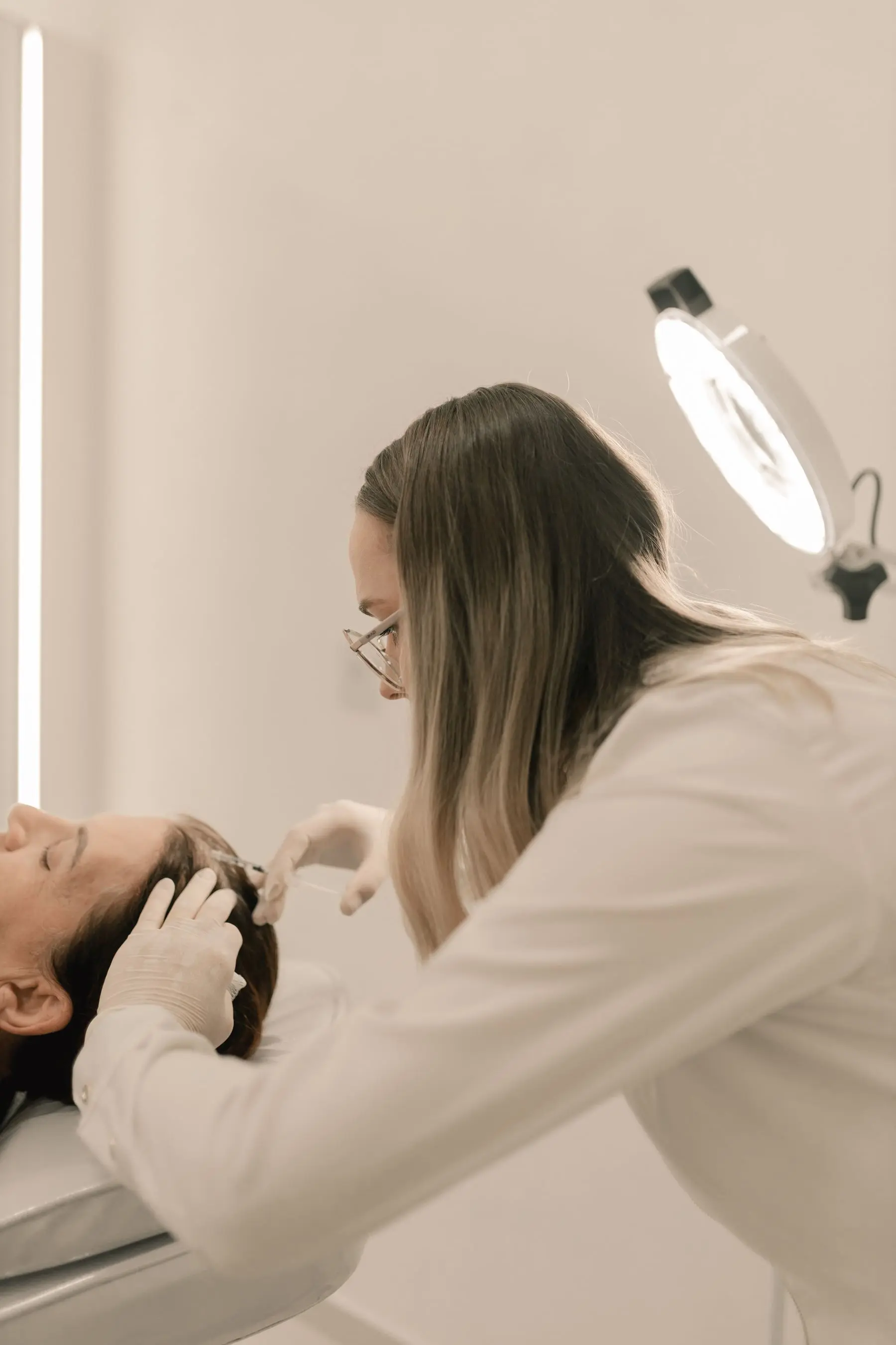 A dermatologist in a cream coat examines a patient's skin under a ring light.