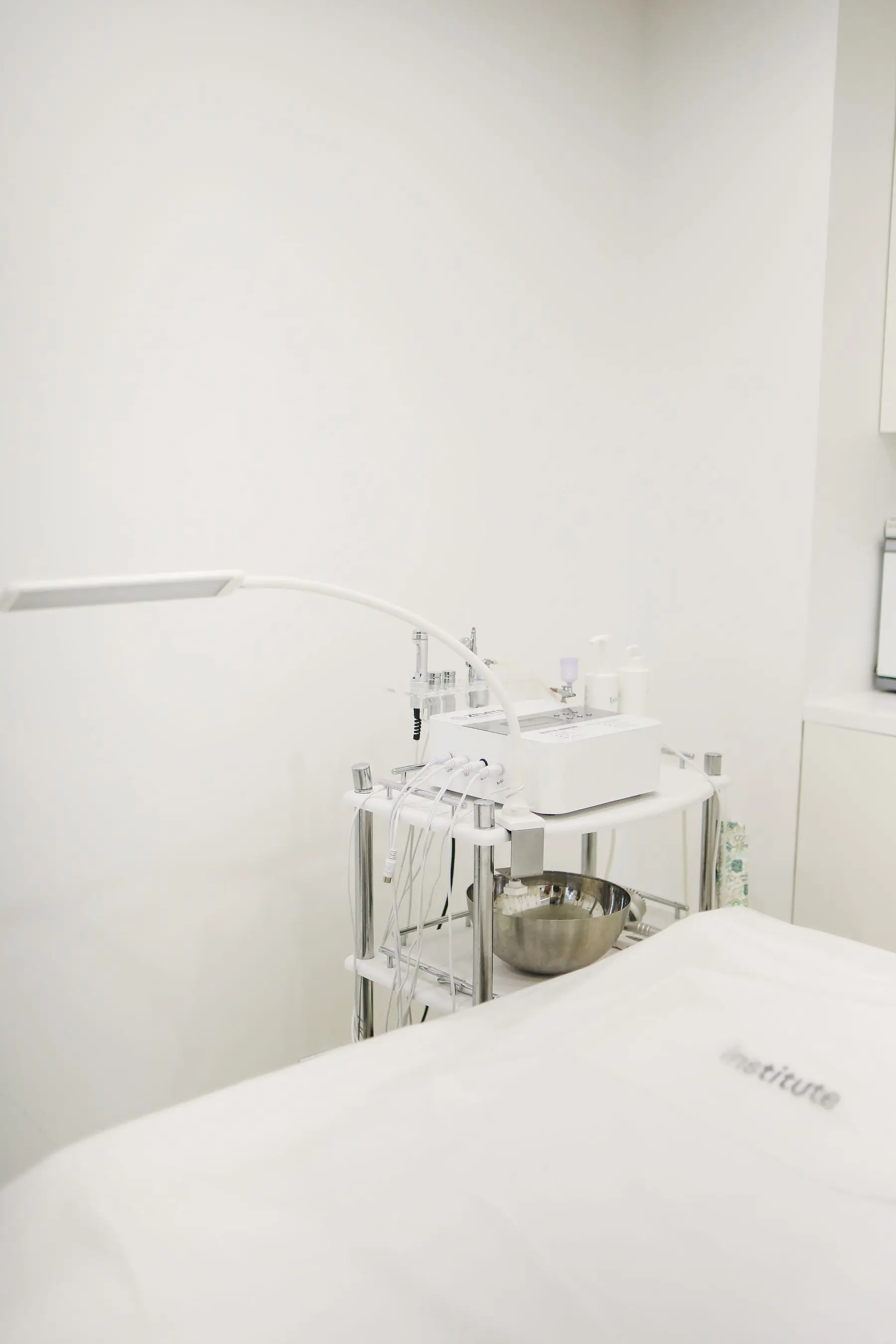 A calm, bright treatment room with a skincare device on a trolley beside a bed.