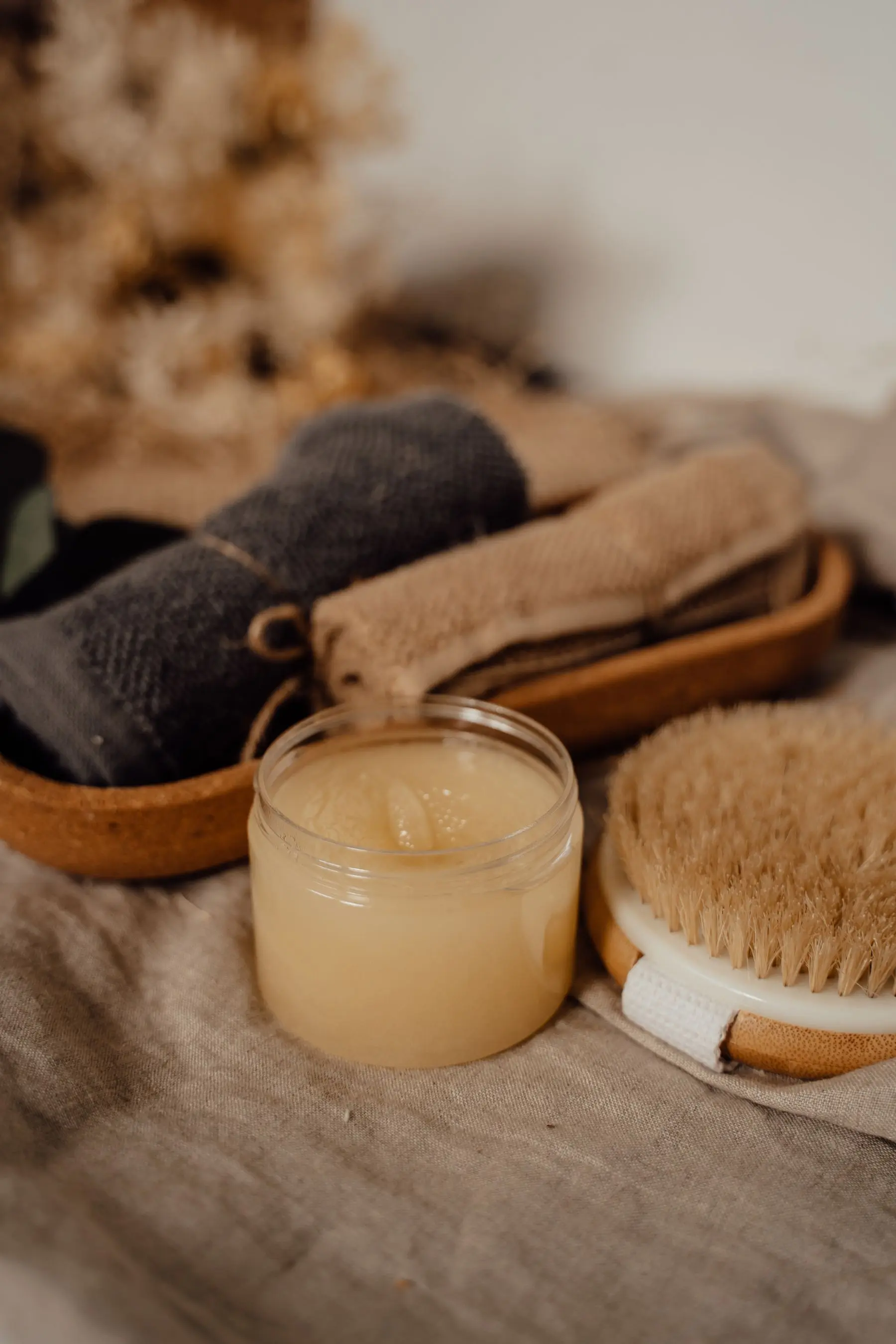 A jar of body scrub with a bristle brush and a folded towel on a woven tray.