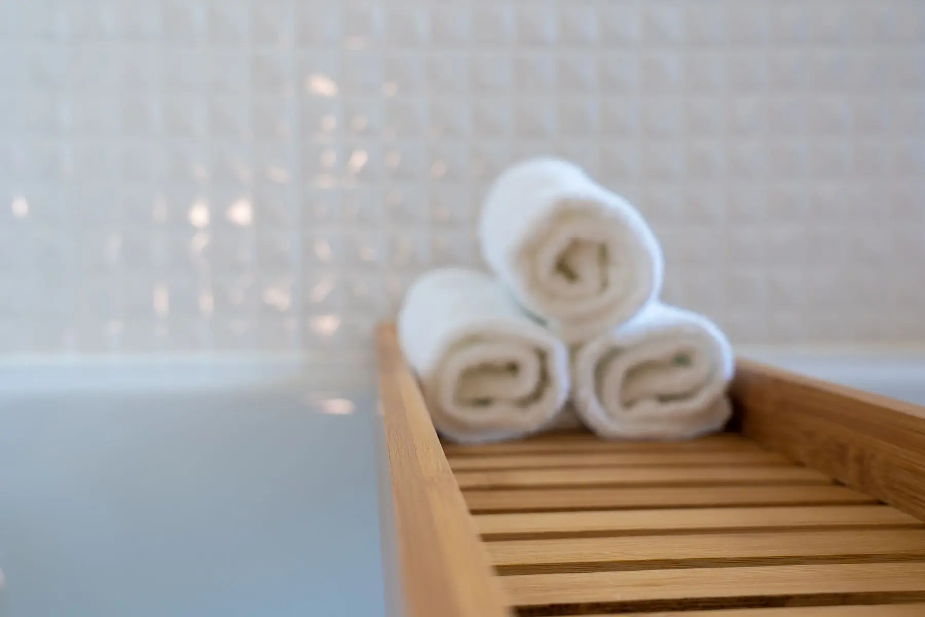 Rolled white towels on a wooden bath tray against white tile.