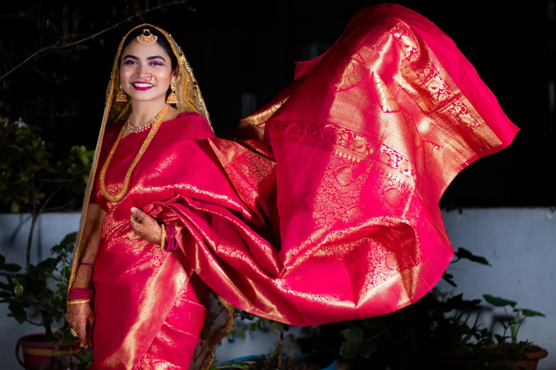 A smiling woman in a bright pink and gold silk saree, the end of the saree billowing behind her.