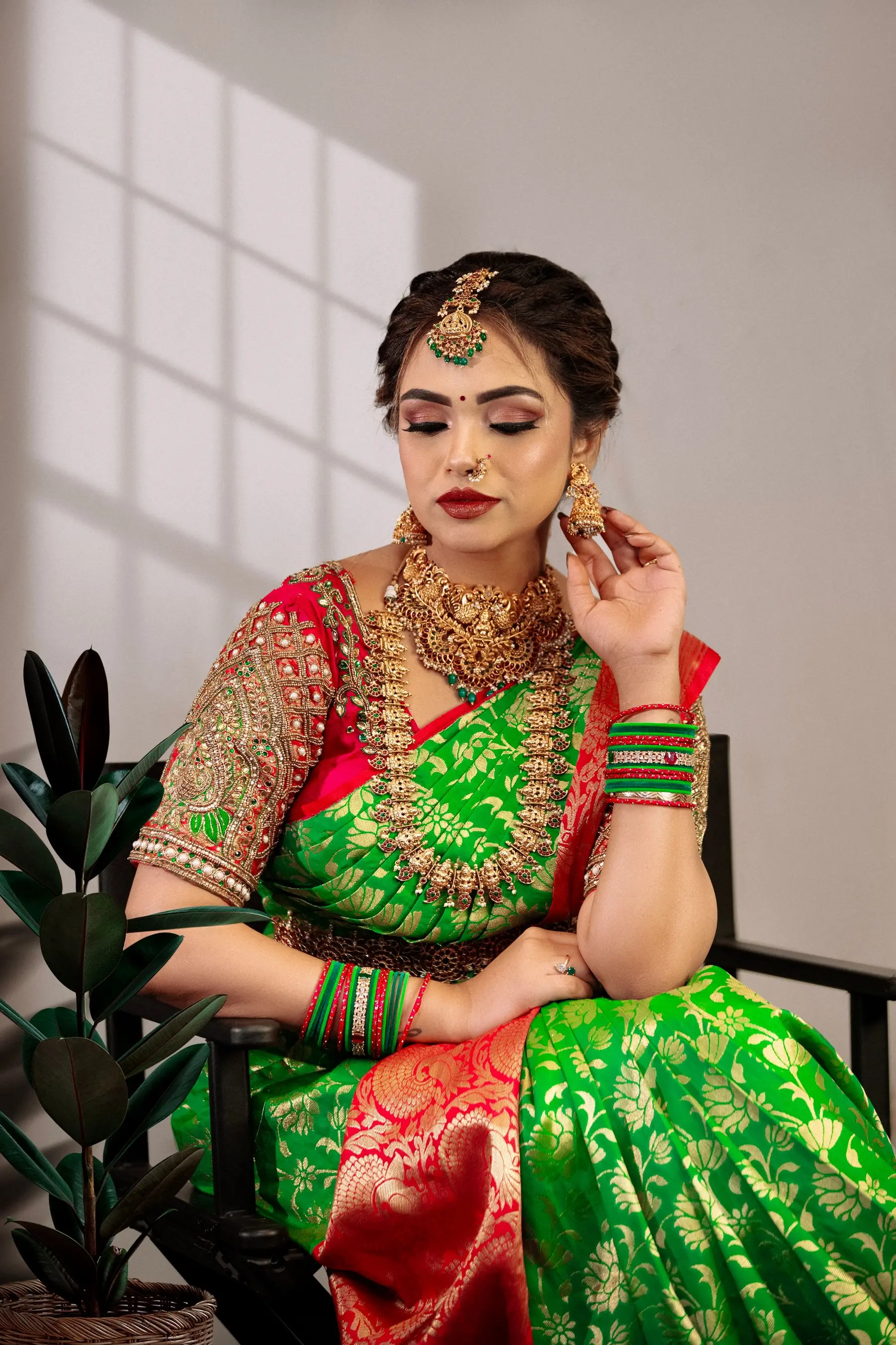 A woman in a green silk saree and red blouse sits with heavy gold necklaces, bangles and a forehead ornament.