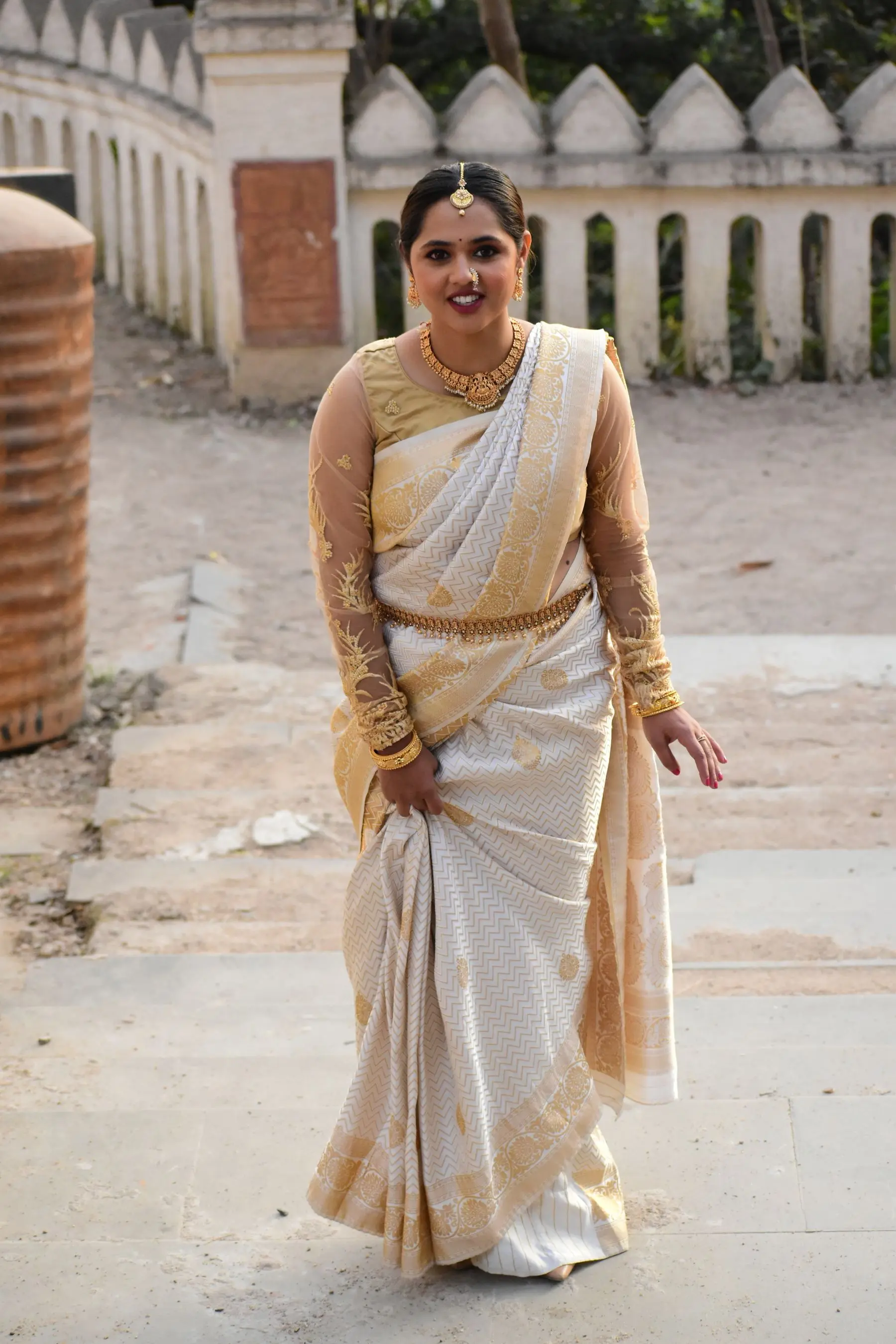 A woman in a white saree with a gold border and a gold waist belt smiles on stone steps.