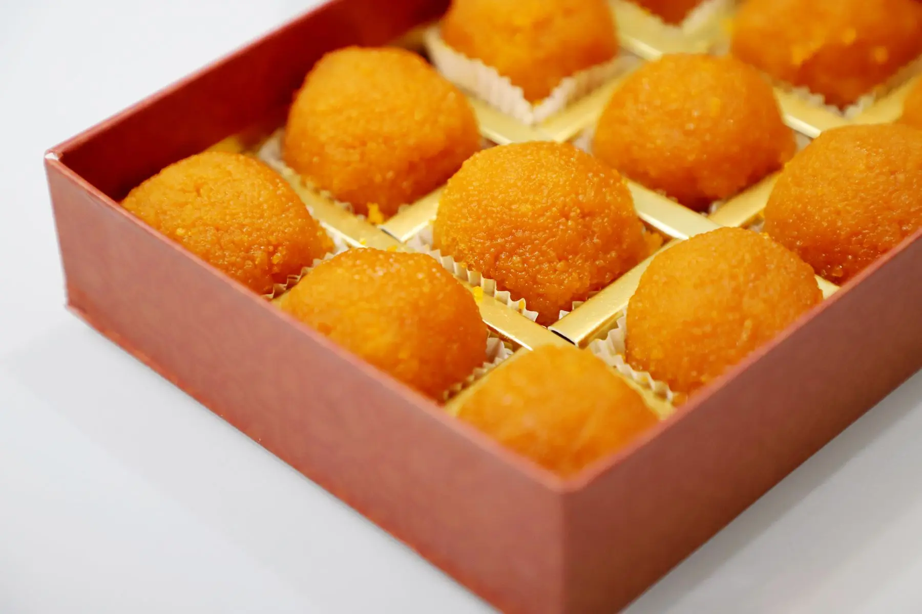 A pink gift box of round orange laddoos, each in a gold paper cup.