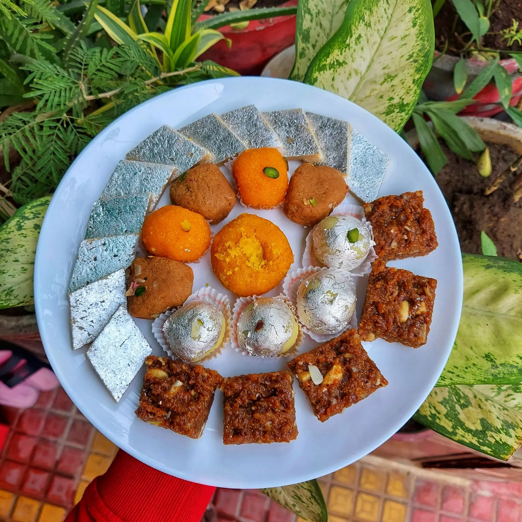 An overhead view of a white platter of assorted barfi, laddoos and silver-leaf sweets on green leaves.