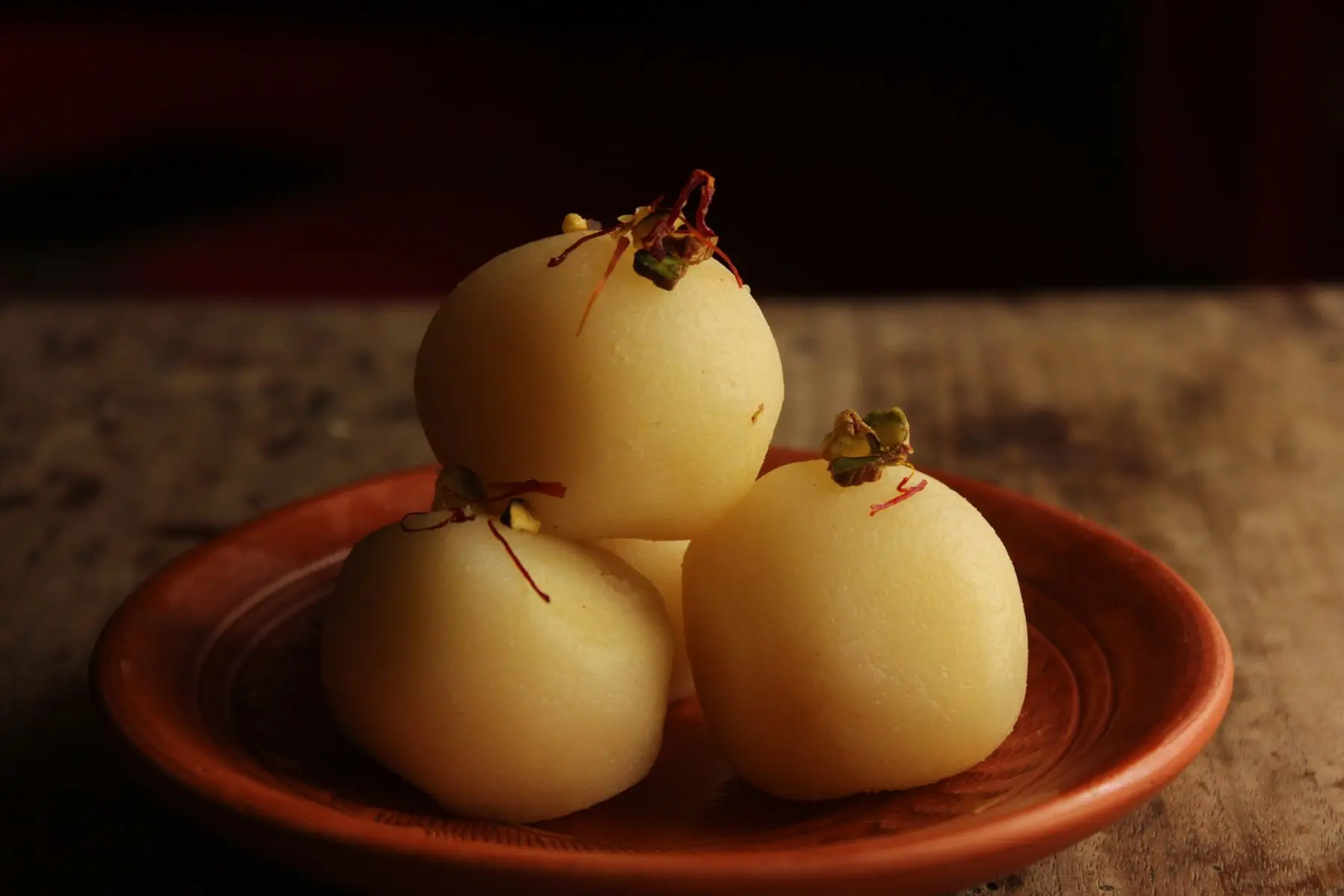 Three pale rasgullas with threads of saffron on a clay plate.