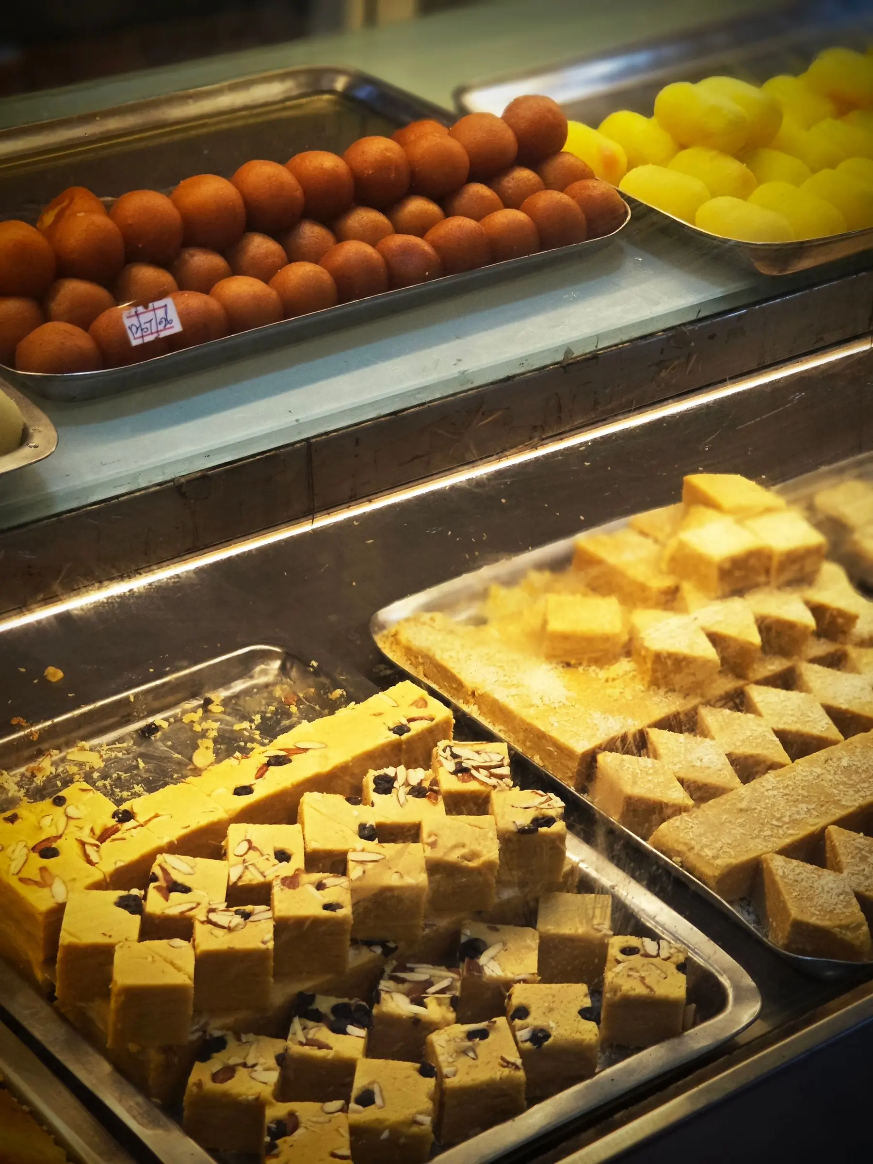 Trays of golden sweets cut into diamonds in a shop display case, with dark gulab jamun above.