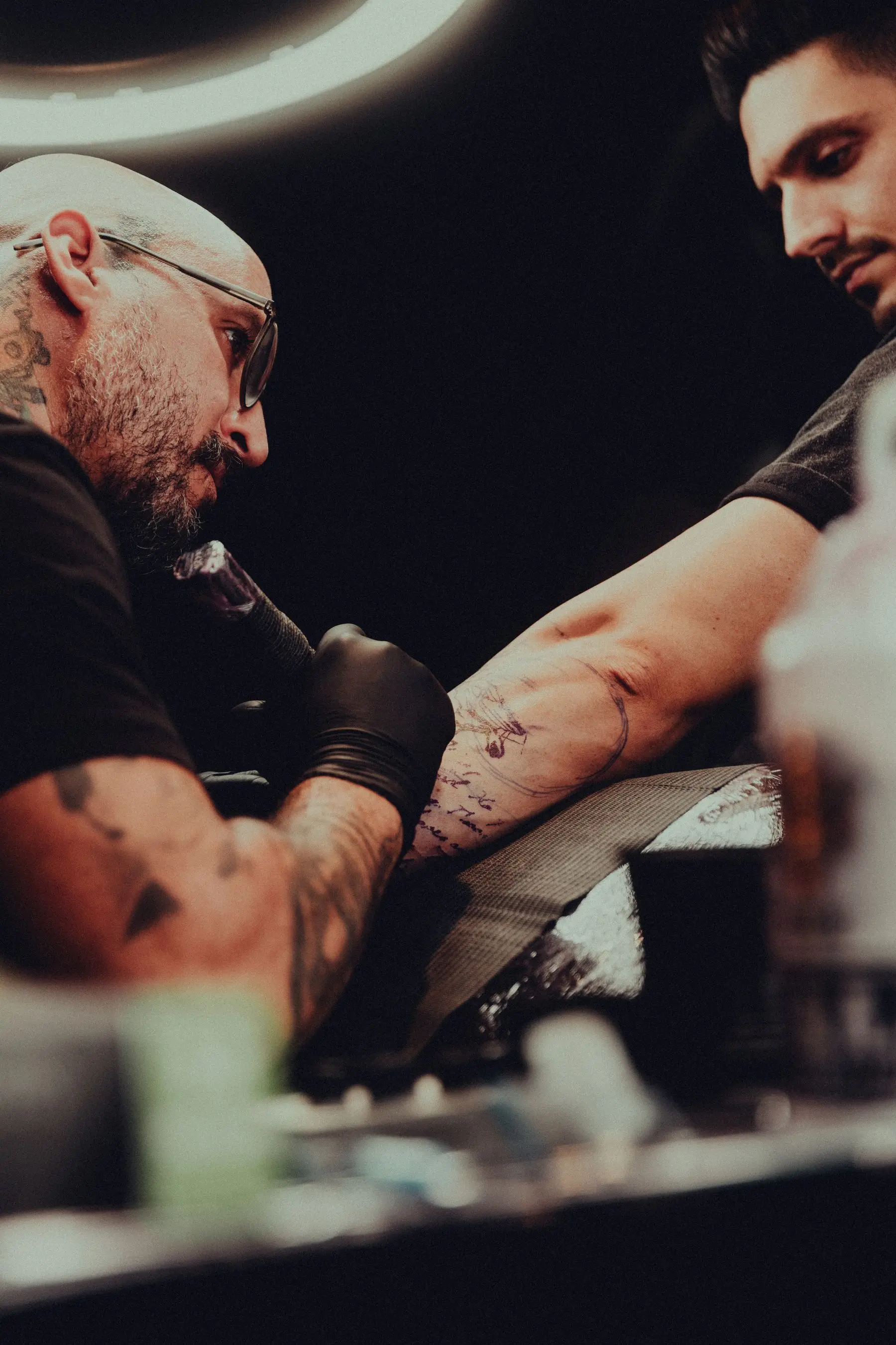 A tattooist in glasses works on a client's forearm, lit in deep amber.