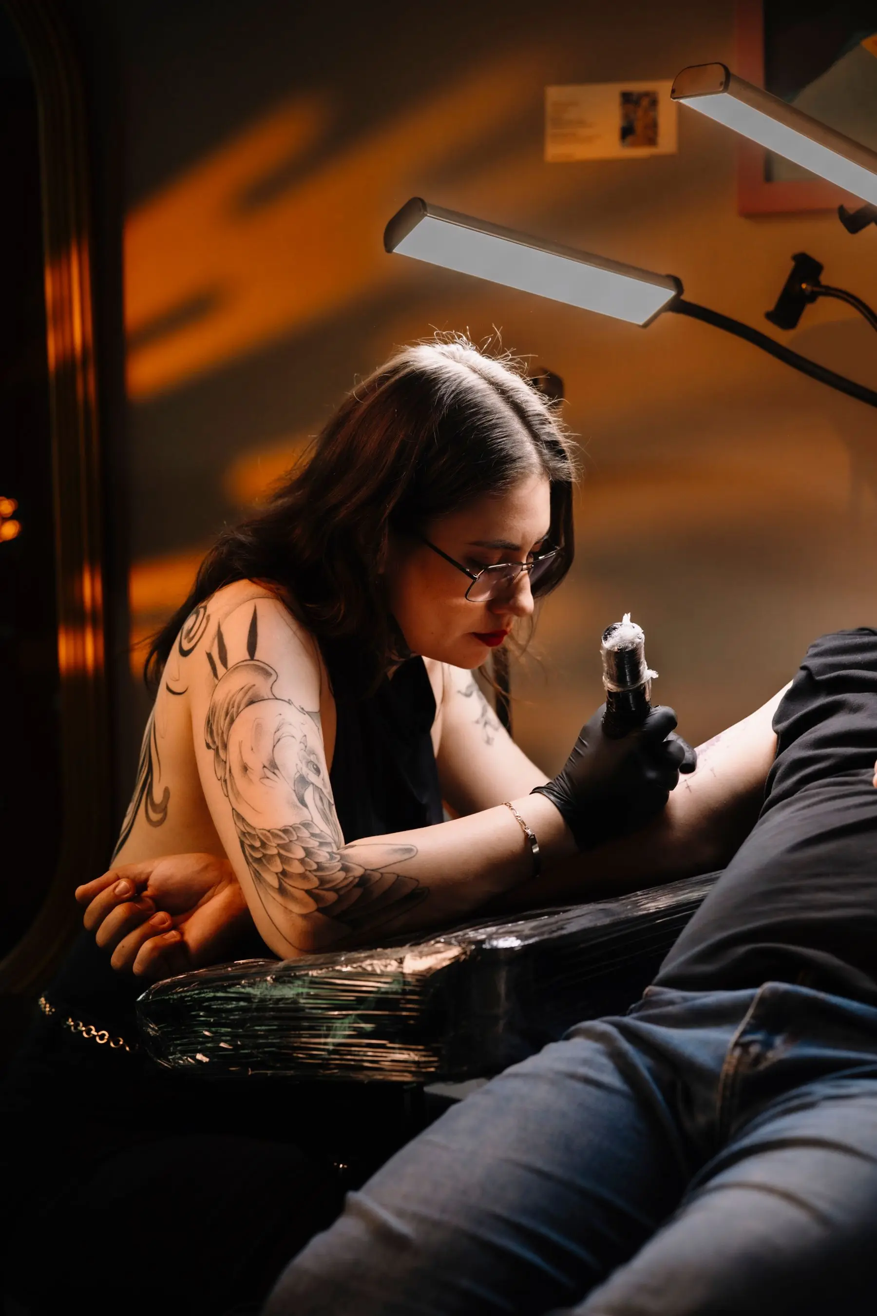A woman artist with a patterned sleeve tattoos a client's arm under a warm lamp.
