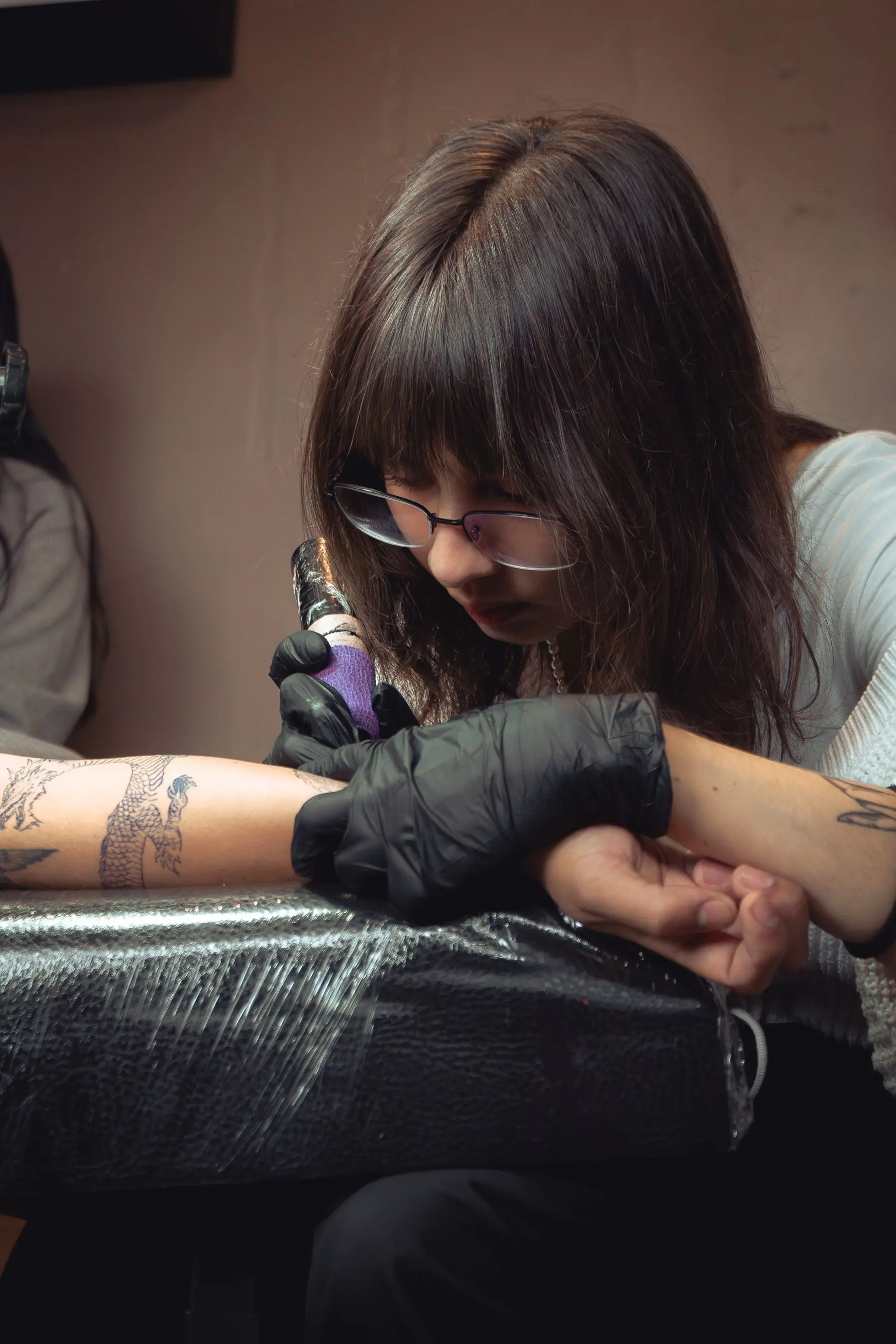 A woman with a fringe leans over a client's arm, a tattoo machine in her gloved hand.