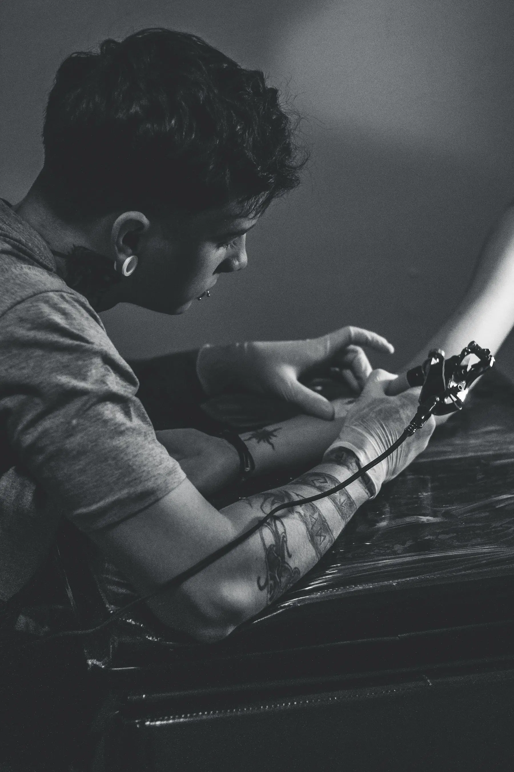 A black-and-white close view of a tattoo machine at work on a forearm.