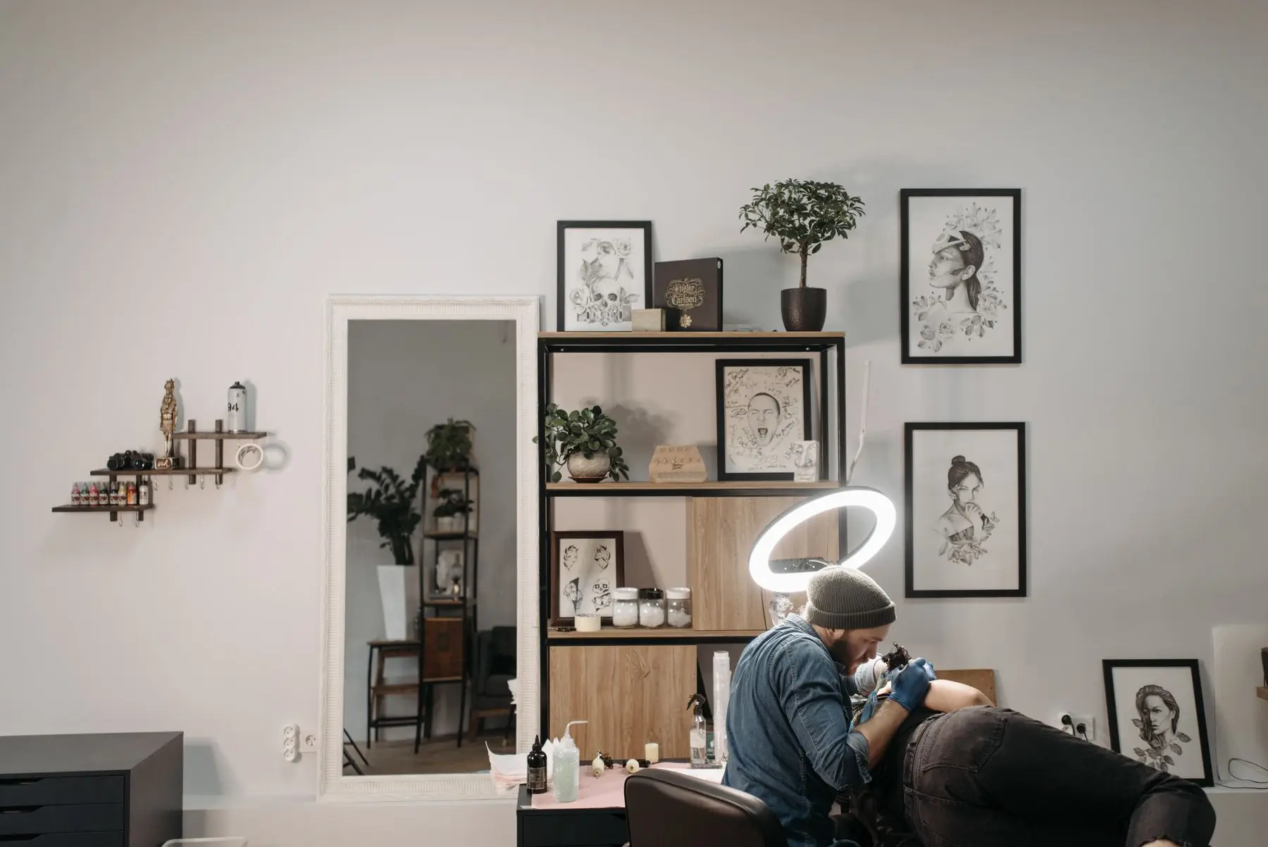 An artist in a beanie works under a ring light in a bright studio with framed flash art and a potted plant on a shelf.