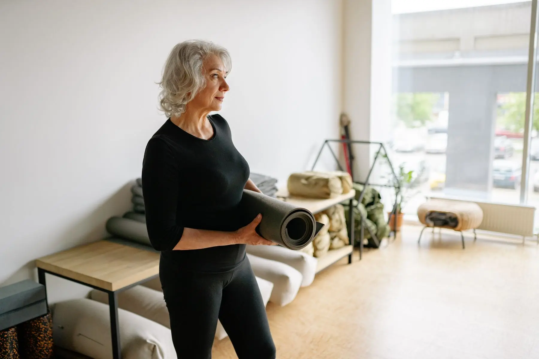 An older woman in black carries a rolled mat in a bright room with cushions and blankets.