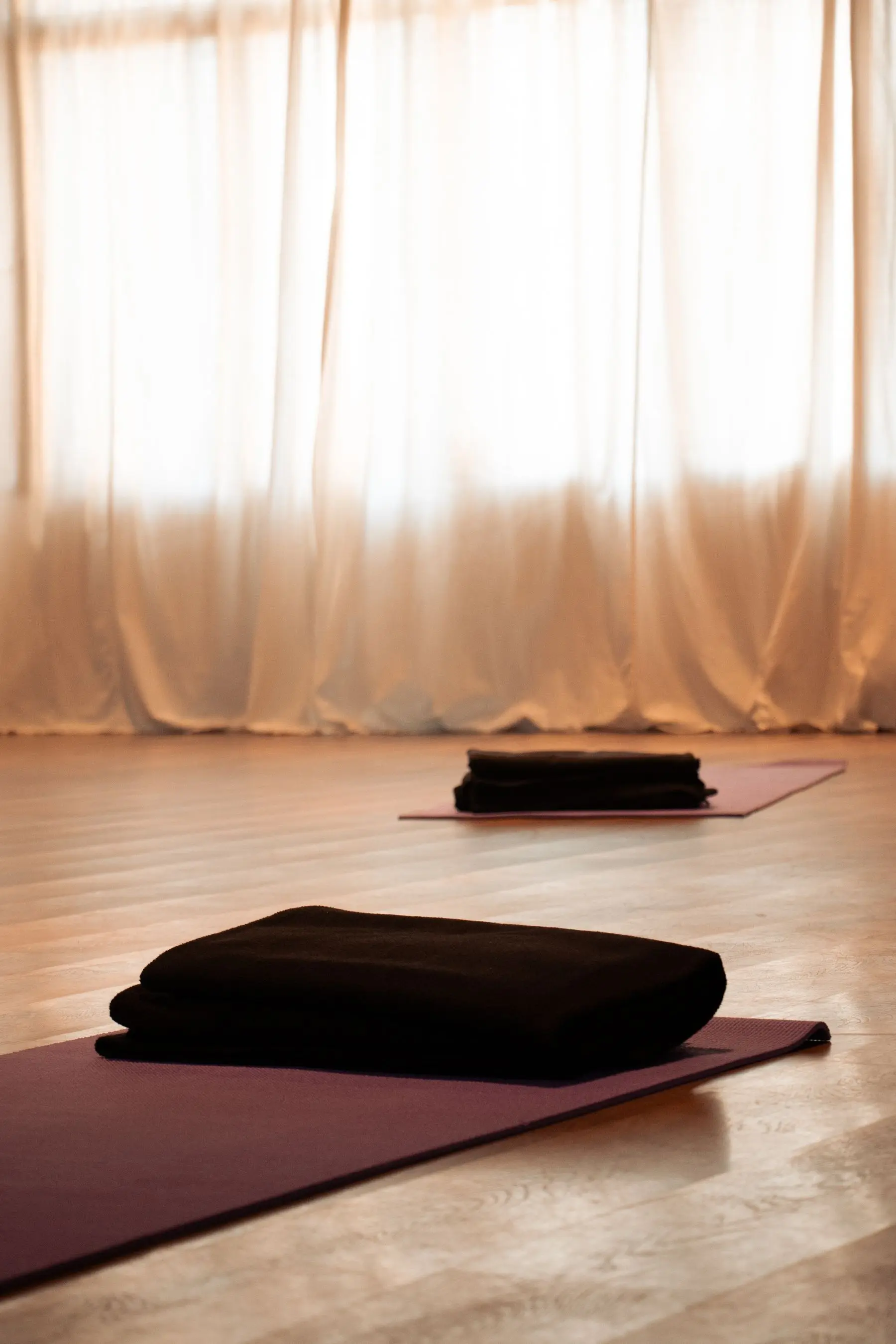 A folded blanket and a mat on a wooden studio floor in front of sheer, sunlit curtains.