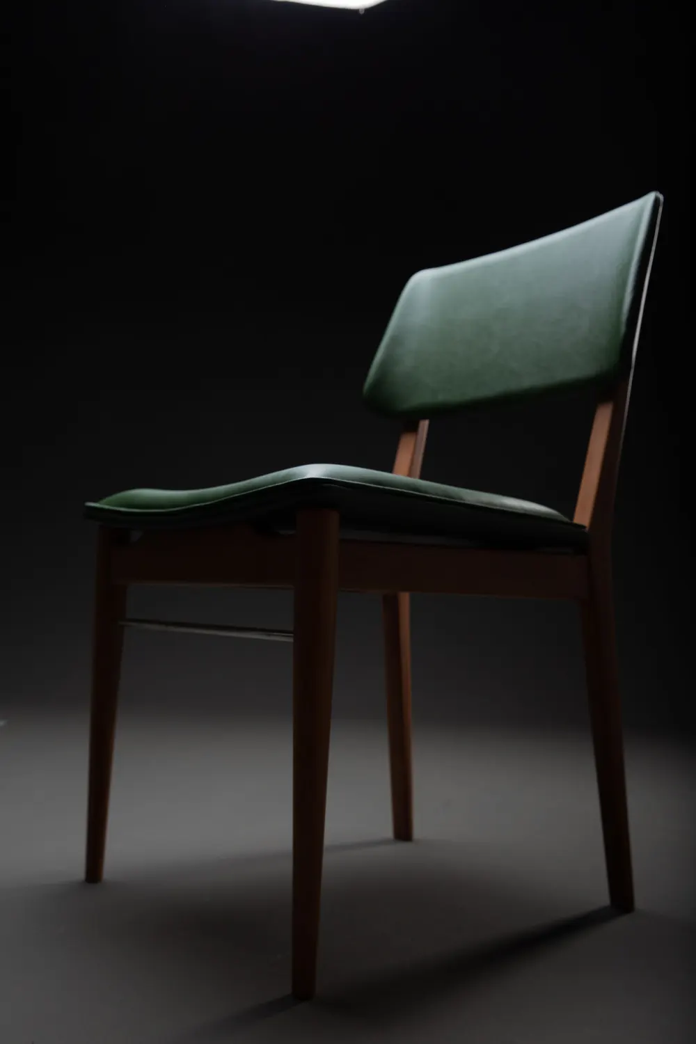 A side chair with a walnut frame and green leather seat and back, lit by one soft light against a dark ground