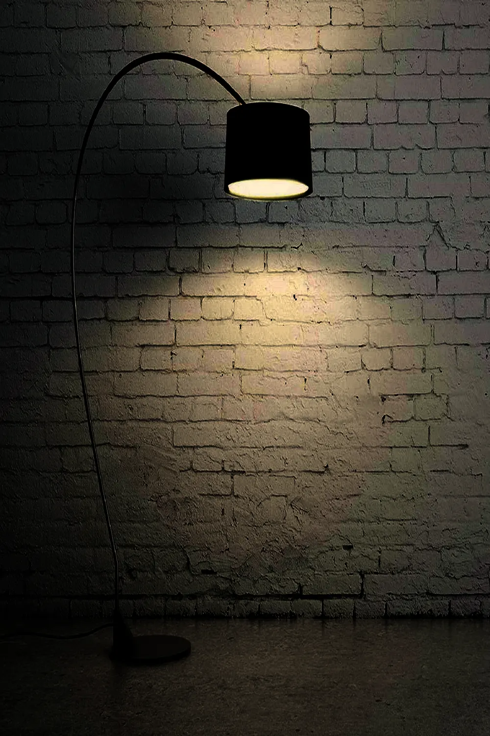 A black arc floor lamp reaching over from the left, its drum shade throwing a pool of light on a dark brick wall