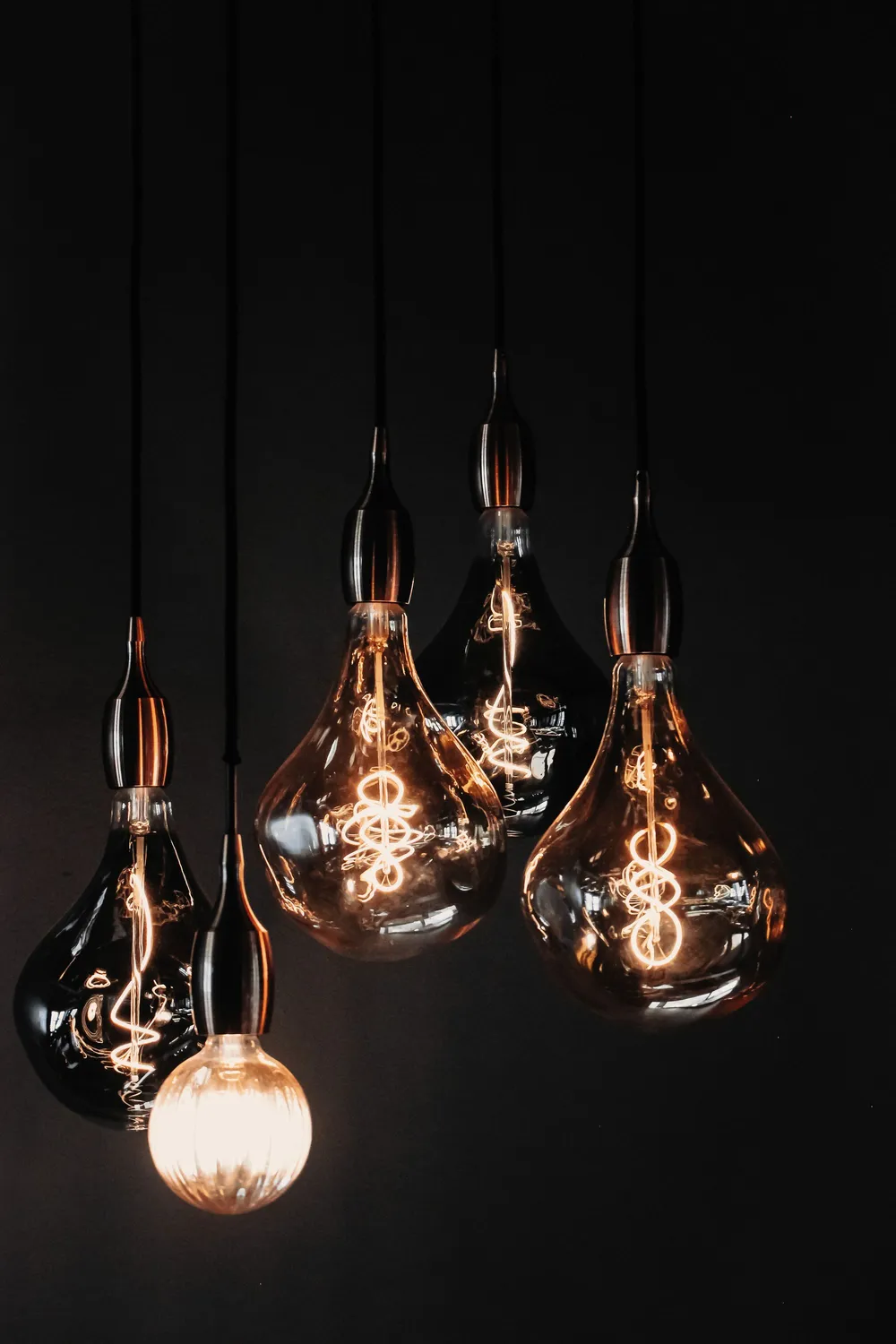 Five glass drop pendant lamps hanging on black cords in the dark, each with a lit filament